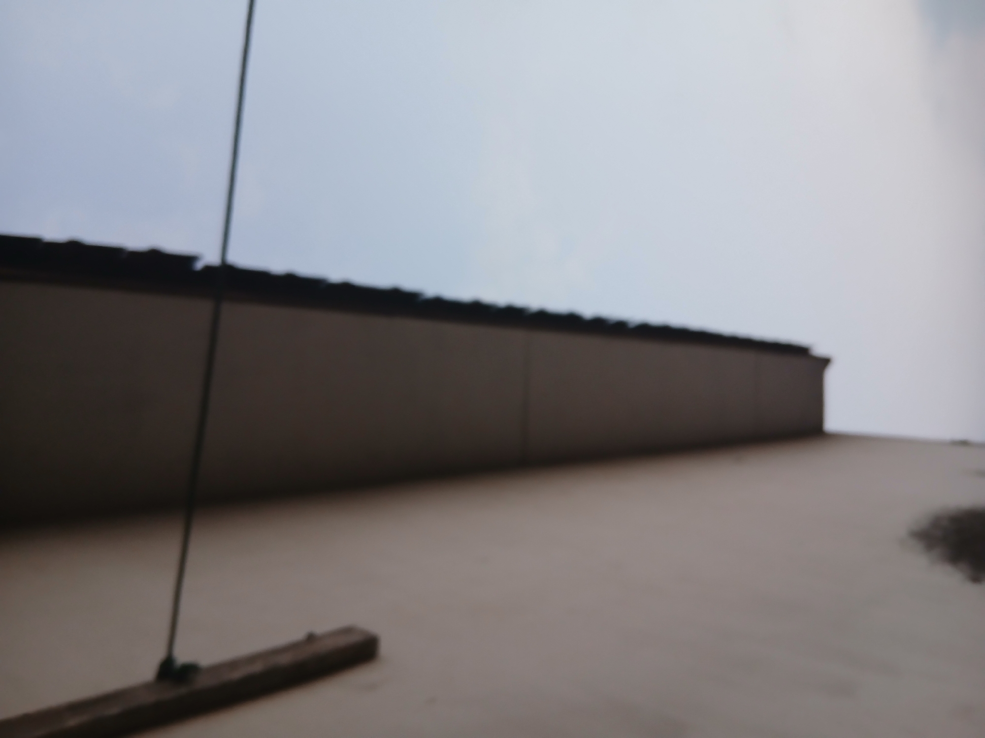 The image shows a side view of a building's eave and wall, with a pale blue sky in the background. A horizontal dark line, likely a wire or cable, stretches across the image, attached to a wooden plank on the wall. The building's wall is a light color, and the eave is dark. The image appears to have been taken from a low angle. There is no discernible location information within the image itself.