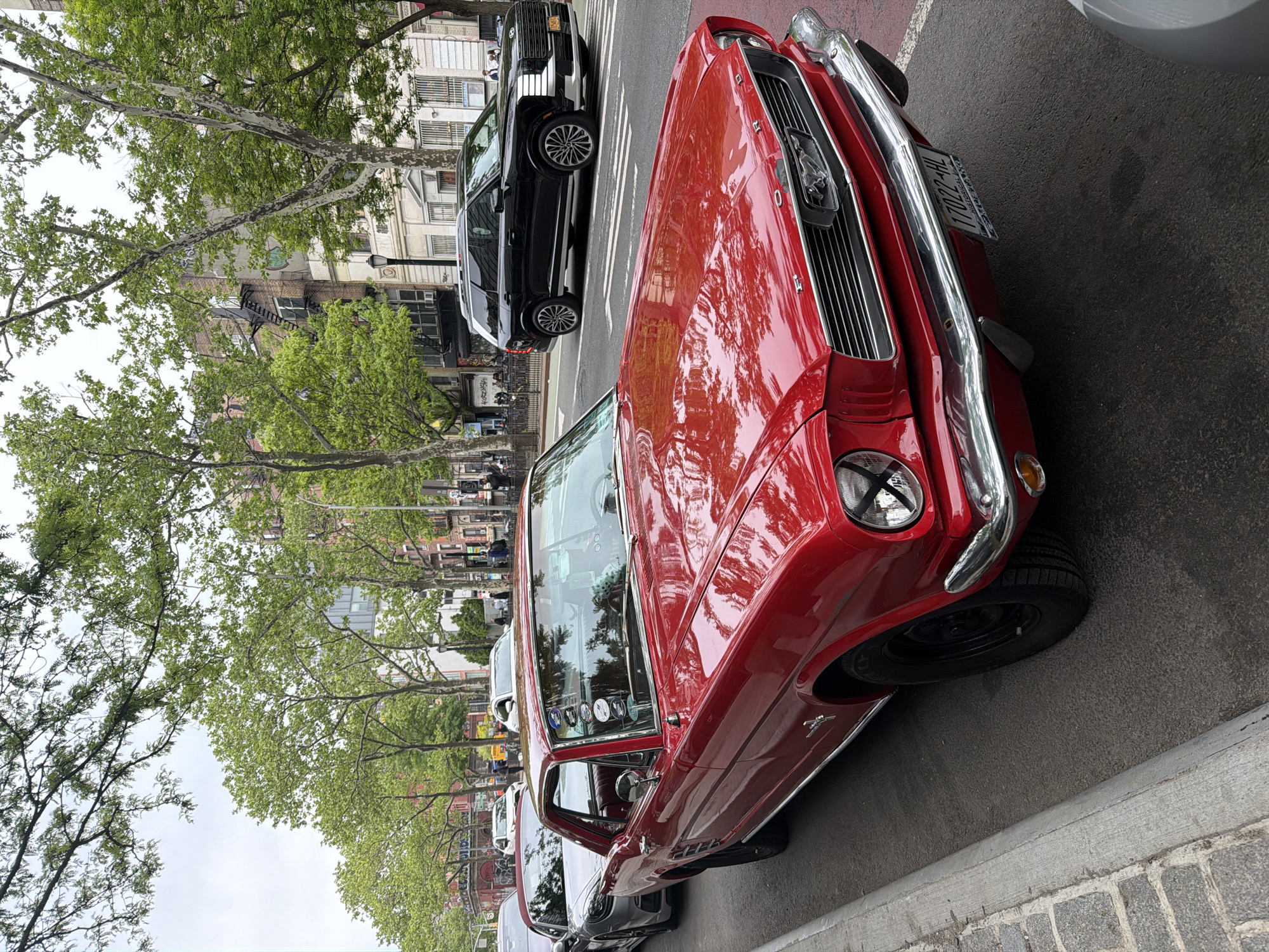 The image depicts a red vintage Ford Mustang parked on a street in New York City, United States of America. The car is positioned along the curb, with other parked vehicles visible behind it. The background reveals buildings typical of New York City, including brownstones and apartment buildings. Trees with green foliage line the street, suggesting it is a residential area. The sky is overcast. A black modern SUV is parked next to the Mustang. The scene appears to be a typical urban street scene with a classic car prominently featured.
