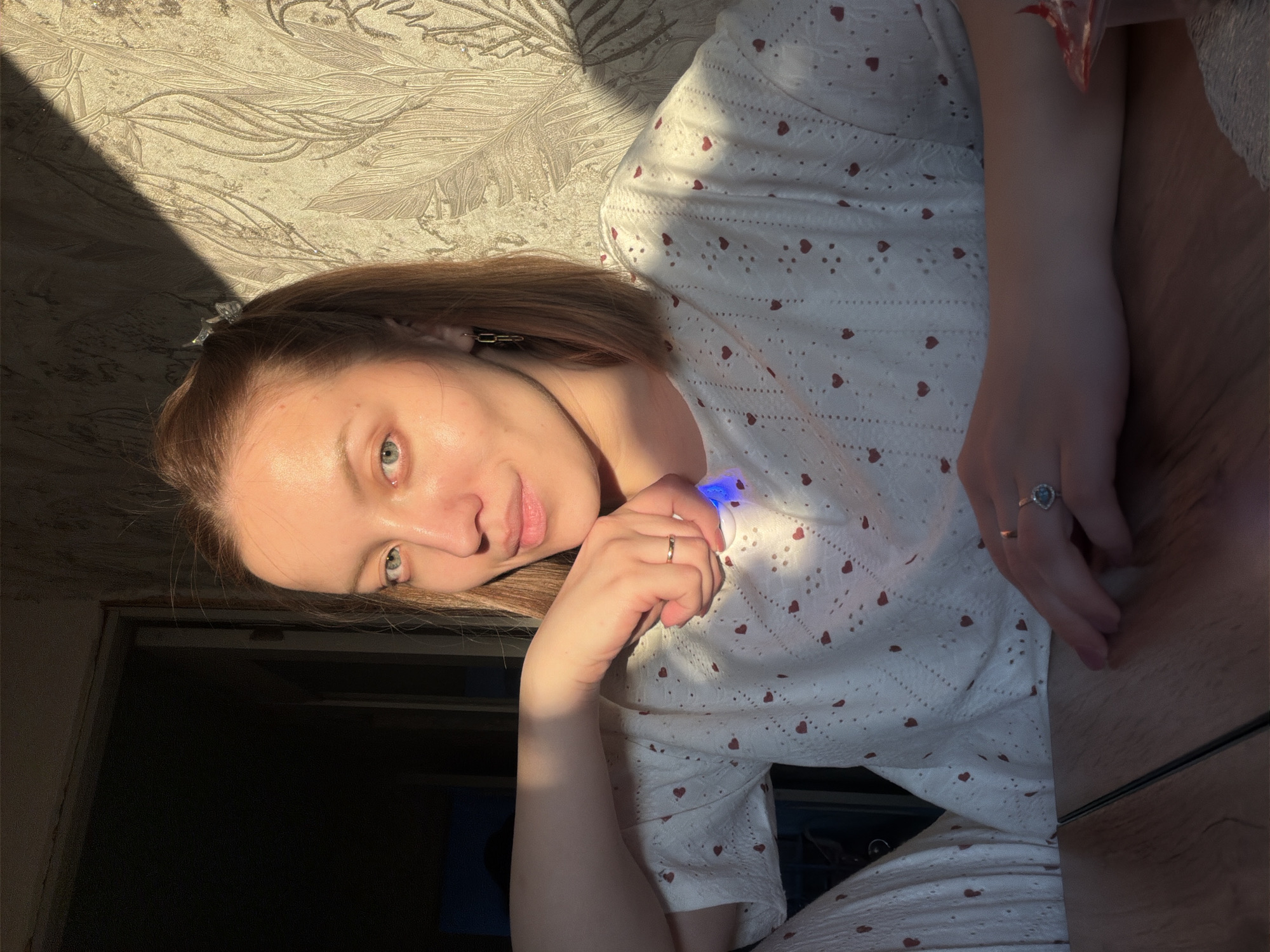 A fair-skinned woman with light brown hair and green eyes is captured in a close-up shot. She is wearing a white top with red heart patterns. She is illuminated by sunlight, with a sharp contrast between the bright areas on her face and clothing, and deep shadows in the background. She holds a small, white, round object with a blue light emitting from it. Her hand has two rings. The background includes a wall with a floral pattern. The photograph appears to be taken in an indoor environment, possibly a room, with portions of a door visible in the lower-left corner. There is insufficient information to determine the exact location in Reshetnikovo, Russia.