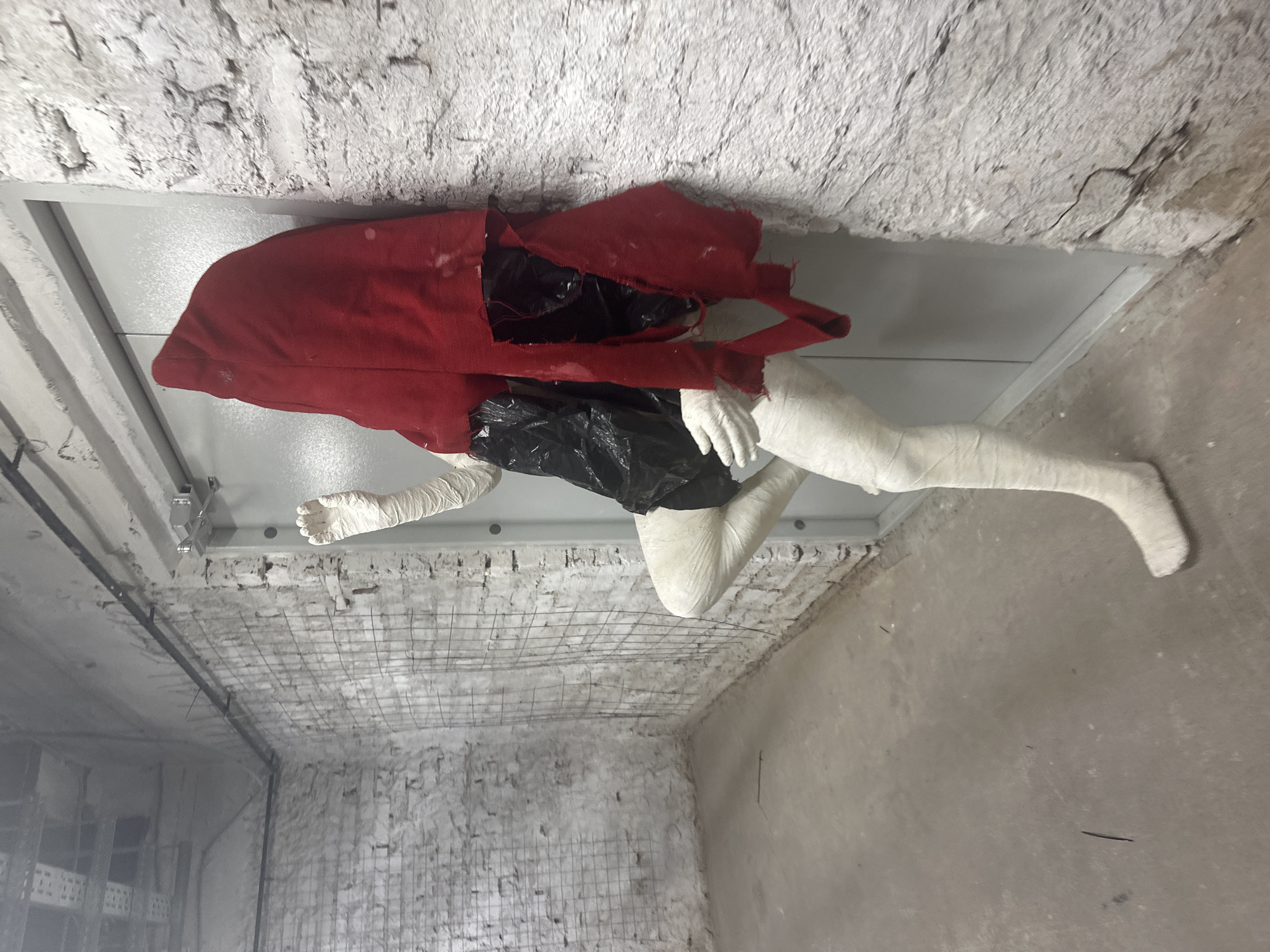 The image shows a partially visible human form that appears to be a mannequin or a sculpture, lying on its side. The form is covered with white bandages, suggesting an injury or a cast. A red cloth is draped over its torso, and a black plastic bag is visible underneath. The background is a rough, unfinished concrete room with exposed rebar on the ceiling, indicating a construction site or an abandoned space. The lighting is artificial and somewhat dim. The scene has an unsettling and possibly artistic quality, evoking a sense of vulnerability or decay. The image is oriented vertically, with the top of the scene appearing on the left side. There is no visible text in the image. The location context suggests it is in Novosibirsk, Russia.