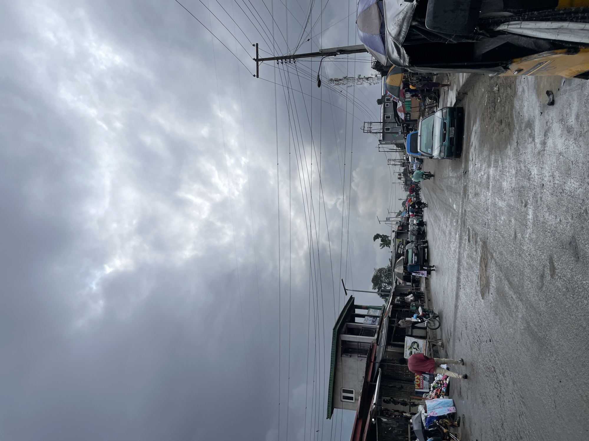 The image depicts a street scene in Ado, Nigeria, under a heavily overcast sky. The road surface is wet and appears to be in poor condition, with puddles and debris visible. Along the sides of the road are makeshift stalls and shops, some with awnings and displays of goods. A few vehicles, including cars, a motorcycle, and a three-wheeled auto-rickshaw, are present on the road. Several individuals are seen walking or standing near the shops. Overhead, numerous electrical wires crisscross the sky, supported by utility poles. The overall atmosphere suggests a busy, albeit somewhat gritty, urban environment.