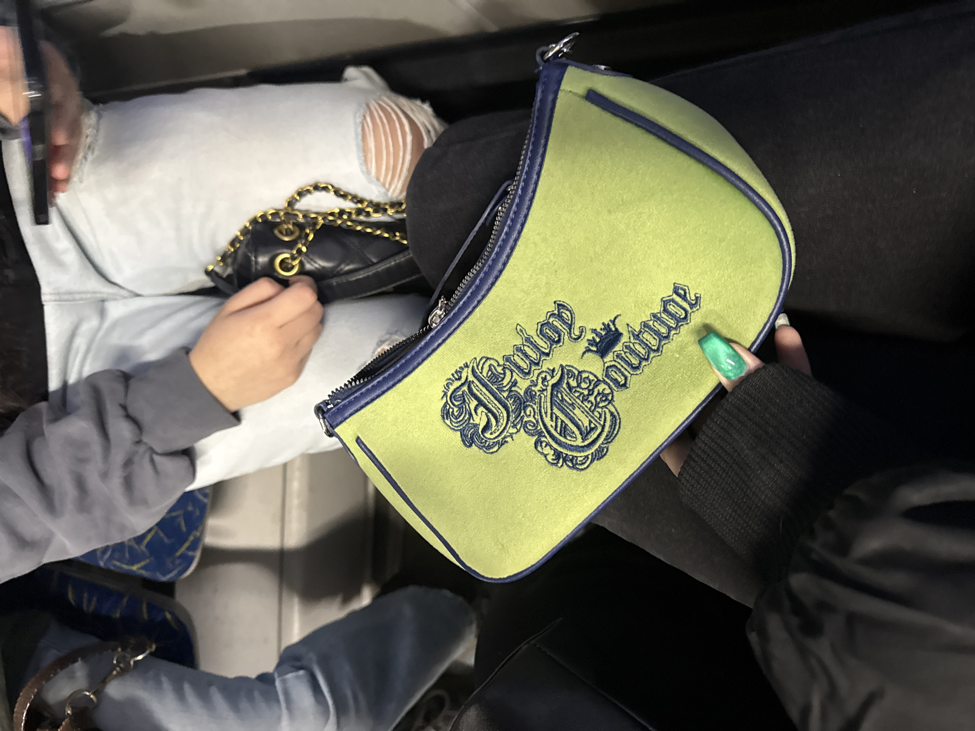 A medium shot from above shows two seated people inside an enclosed space, likely public transportation. The first person, on the left, is wearing distressed light blue jeans and a gray sweatshirt. They hold a mobile phone in their left hand. A small black quilted purse with a gold chain is on their lap. The second person, on the right, wears dark-colored clothing and holds a light green "Juicy Couture" purse with dark blue embroidery and trim. Their nails are painted green. The interior has blue seat covers with yellow patterns. There is no visual information that suggests this image was taken in Saint Petersburg, Russia.