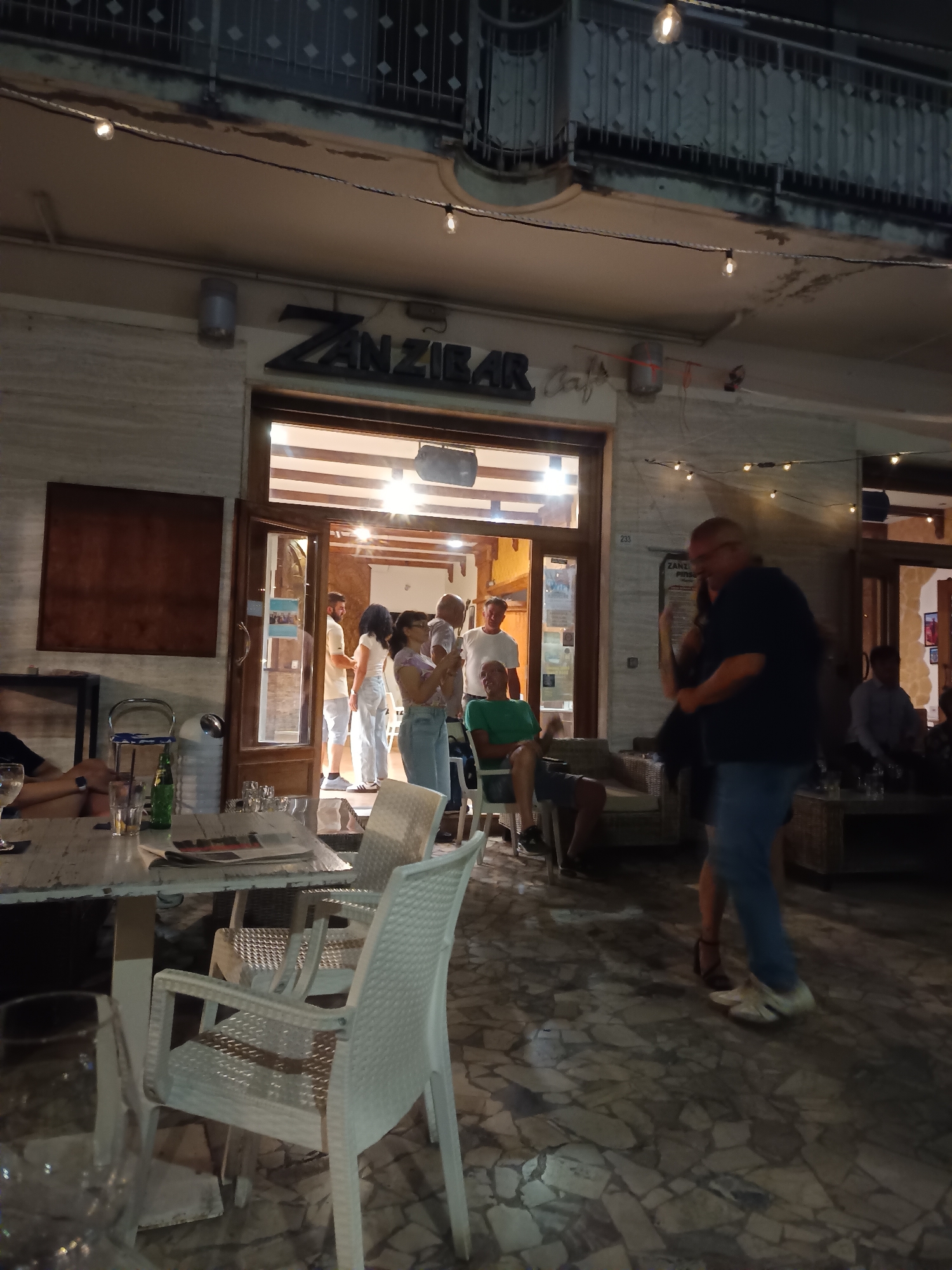 The image depicts an outdoor seating area in front of a establishment identified by the signage "ZANZIBAR cafe" in Pineto, Italy. It appears to be nighttime, with string lights illuminating the scene. Several people are present. In the foreground, white plastic chairs surround a table with drinks and newspapers. Further back, a couple is dancing, with the male figure wearing a dark blue shirt and jeans, and the female figure in a dark dress. Other individuals are visible standing or sitting near the entrance of the cafe. The ground is paved with a mosaic of stones. The architecture suggests a Mediterranean style.
