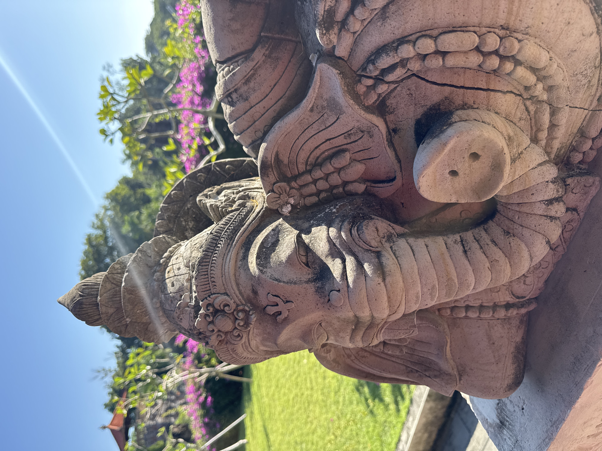 A stone statue of the Hindu deity Ganesha, depicted in a reclining pose, dominates the foreground. The statue, appearing weathered and light brown-grey, features intricate carvings on its elephant head, crown, and body. Its trunk is depicted in a simplified, circular form with two nostril-like holes. The statue rests on a light-colored stone base, which is partially visible in the lower foreground.
The background reveals a brightly lit outdoor environment under a clear blue sky, with a prominent diagonal sunbeam or lens flare in the upper right quadrant. A manicured green lawn occupies the middle ground on the left. Beyond the lawn and extending across the middle and right background are blurred clusters of vibrant purple-pink flowering bushes, likely bougainvillea, interspersed with various green trees and foliage. A small, distinctive tree with light green leaves is visible centrally in the upper background. In the distant left background, a traditional red-roofed pavilion or gazebo is partially discernible amidst the greenery. The scene is illuminated by strong direct sunlight, indicative of Kuta Selatan, Indonesia.