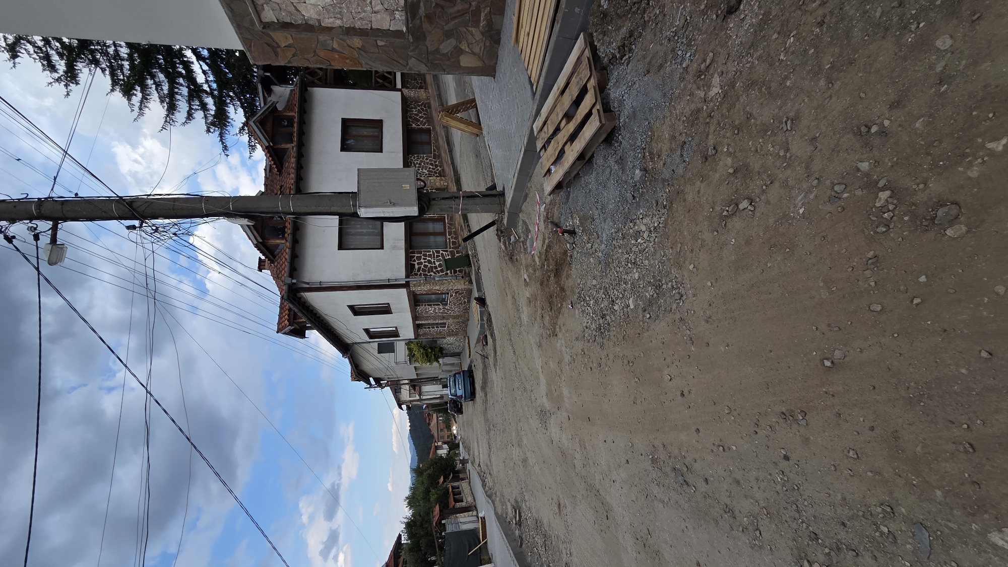 The image depicts a street under construction in the city of Bania, Bulgaria. The street is unpaved and appears to be made of dirt and gravel, with construction materials like wooden pallets and piles of stones visible. Several houses line the street, some with traditional architecture featuring tiled roofs and stone accents. Electrical wires are strung across poles, indicating active utility infrastructure. A blue car is parked on the side of the road, and in the background, a line of more houses and trees can be seen stretching towards a hilly landscape under a cloudy sky. The overall impression is a quiet, rural or semi-rural setting undergoing development.
