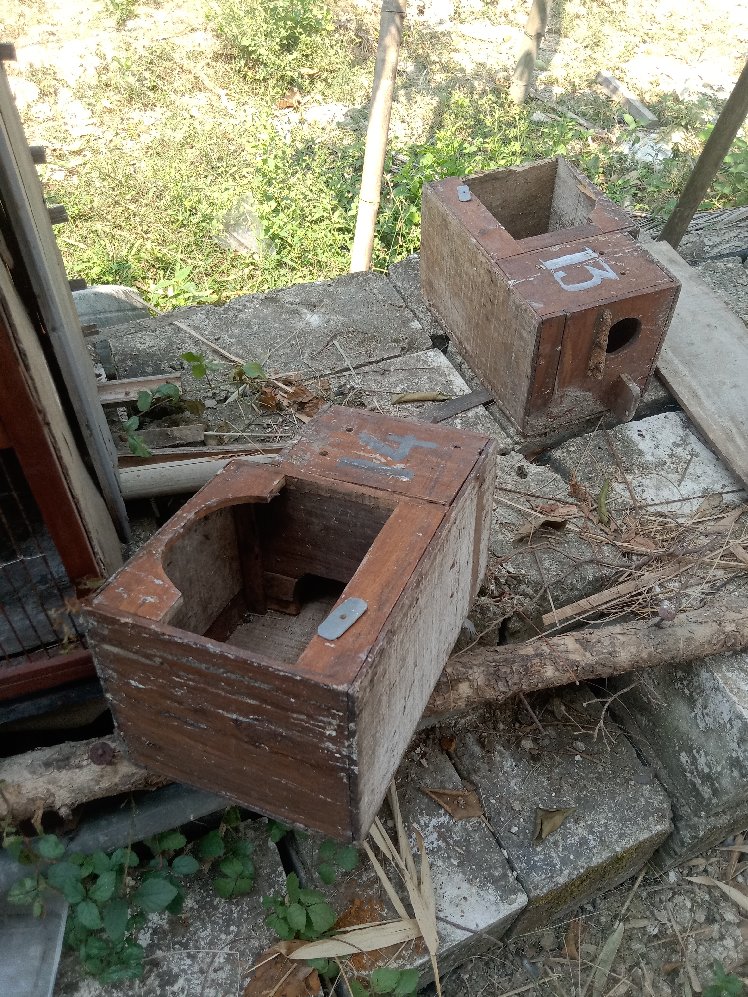 In the city of Stiaji, Indonesia, two weathered wooden birdhouses sit on a rough, uneven surface composed of concrete blocks, debris, and dry foliage. The birdhouses are rectangular with open tops, revealing their hollow interiors. One birdhouse, positioned closer to the foreground, has a metal clasp on its upper edge. The second birdhouse, further back and to the right, is marked with the number "15" in a light color on its side and features a circular opening, indicative of an entrance for birds. Surrounding the birdhouses is sparse vegetation and dry grasses, with a few bamboo stalks visible. The scene appears to be outdoors, under natural daylight.