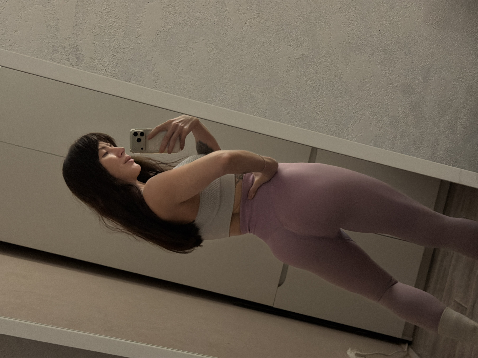 A woman with dark hair is taking a selfie in a mirror. She is wearing a light-colored sports bra and lavender leggings. The background includes a white cabinet and a textured wall. The location in Perm, Russia cannot be confirmed from the image.