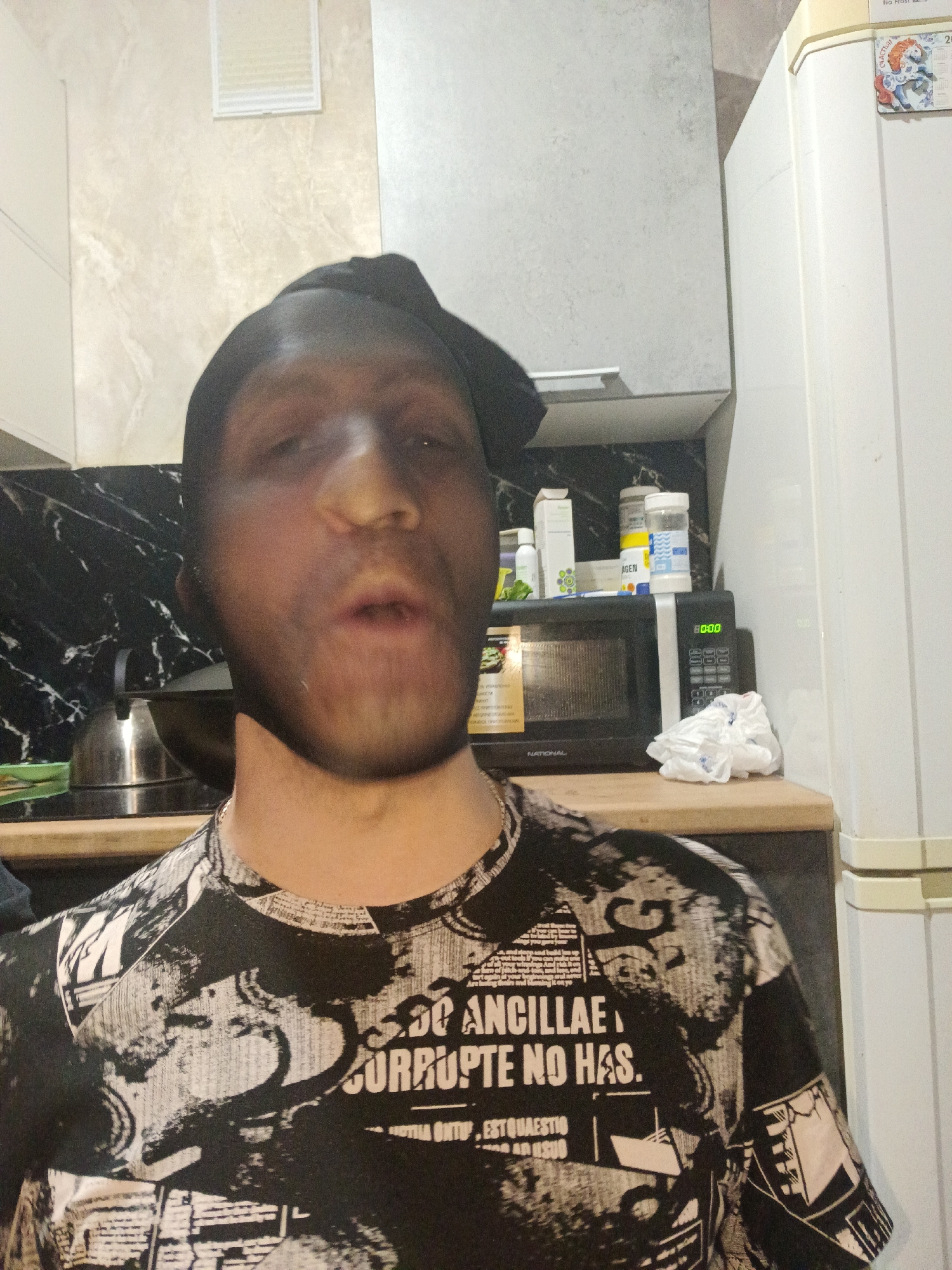 A man in a graphic t-shirt and black head covering is indoors. Behind him are kitchen cabinets, a black microwave, and a white refrigerator. The microwave's display reads "80:00". There is a tea kettle on the counter. Small containers sit on top of the microwave. The man's location is in Chaikovsky, Russia.