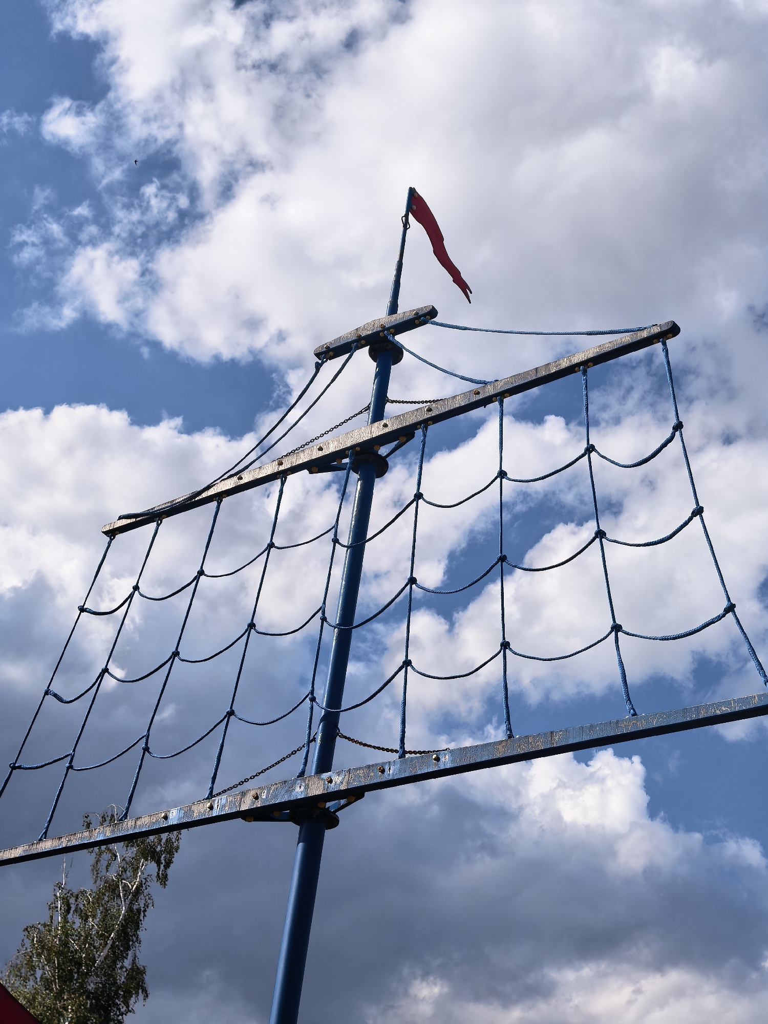 This image features a close-up, low-angle view of a blue mast with a netting structure resembling a sail, set against a cloudy sky. A small red flag flutters from the top of the mast. The netting is made of thick blue rope, woven into a grid pattern and attached to horizontal wooden beams that are bolted to the mast. The wooden beams appear weathered and slightly rusted. In the background, dramatic clouds fill the sky, with patches of blue sky visible between them. A single bird is visible in flight in the upper left portion of the frame. The bottom left of the image shows the upper branches of a birch tree. The overall impression is of a playground structure designed to resemble a ship's mast, possibly located in a park or outdoor recreational area in Sterlitamak, Russia. The lighting suggests it is daytime, with a mix of sun and cloud cover creating a dynamic sky.