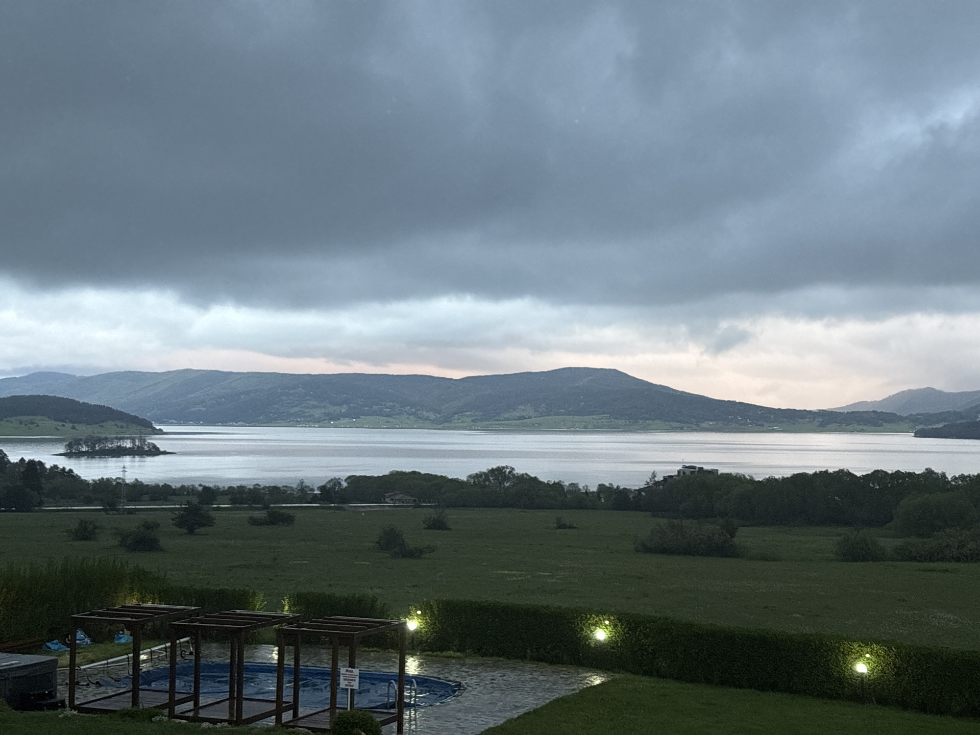 The image depicts a view in Tsigov Chark, Bulgaria, overlooking a lake and mountains under a heavy, overcast sky. The foreground shows a well-maintained lawn with a small swimming pool area covered by wooden frames. The water appears dark and still, suggesting it might be raining or have recently rained. A dense hedge separates the lawn from the pool area. In the mid-ground, a field stretches out towards the lake, with sparse trees and bushes dotting the landscape. The lake fills much of the frame's center, reflecting the grey sky. Mountains rise in the distance, their slopes covered with trees. The overall scene suggests a serene, possibly remote, location.