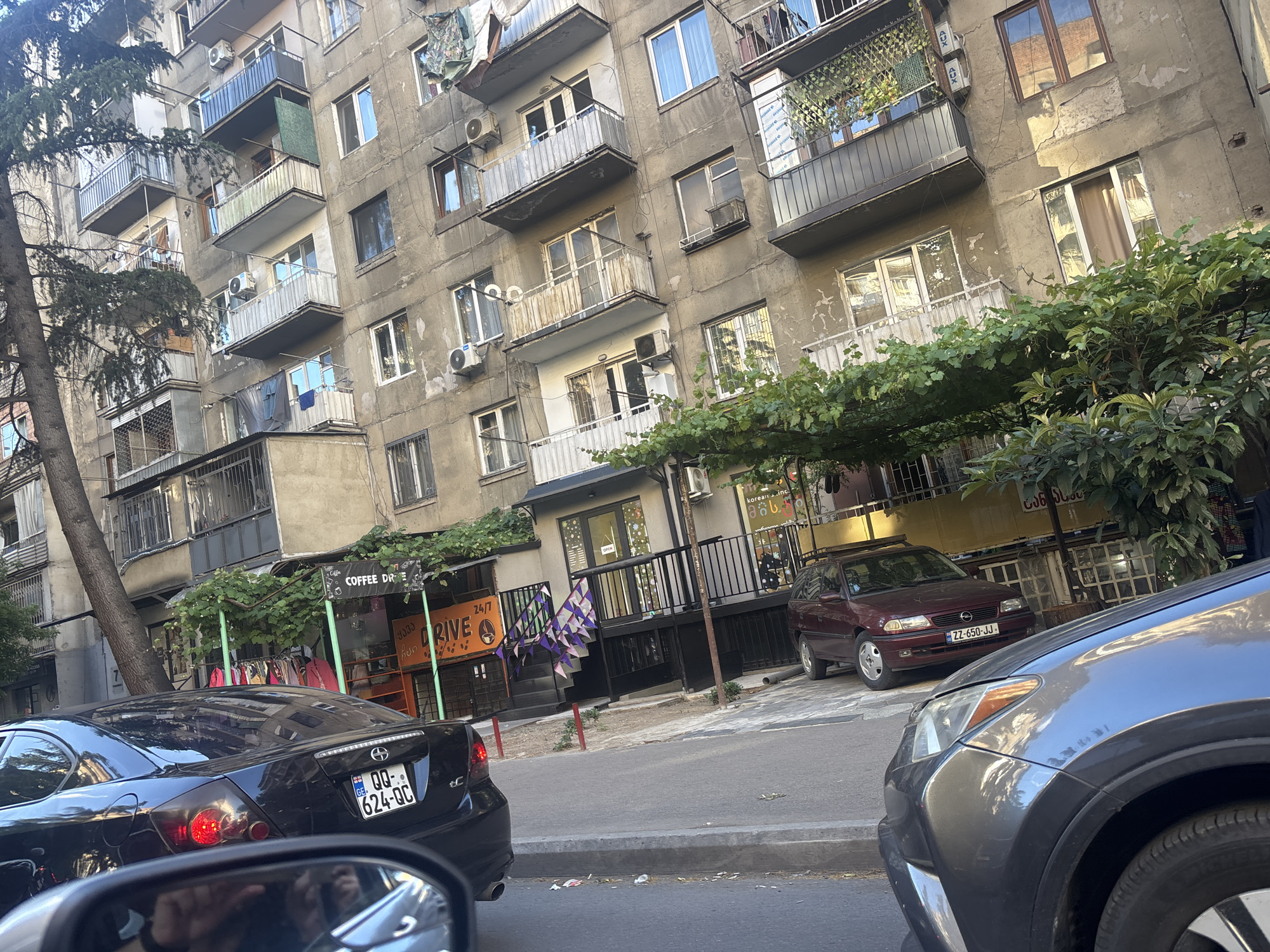 The image captures a street scene in Tbilisi, Georgia, featuring a multi-story apartment building with a weathered facade, numerous balconies with laundry hanging out, and several air conditioning units. In the foreground, two cars are visible: a black sedan with its taillights illuminated and a grey SUV. The black sedan has a license plate that reads "QQ 624-QC". A maroon station wagon is parked further down the street, its license plate showing "ZZ 650-JJ". A building with storefronts is situated at the base of the apartment building, one of which appears to be a "COFFEE DRIVE" establishment. Green vines are growing along a structure in front of the storefronts. The overall impression is a typical urban environment in Tbilisi, with a mix of residential and commercial spaces.