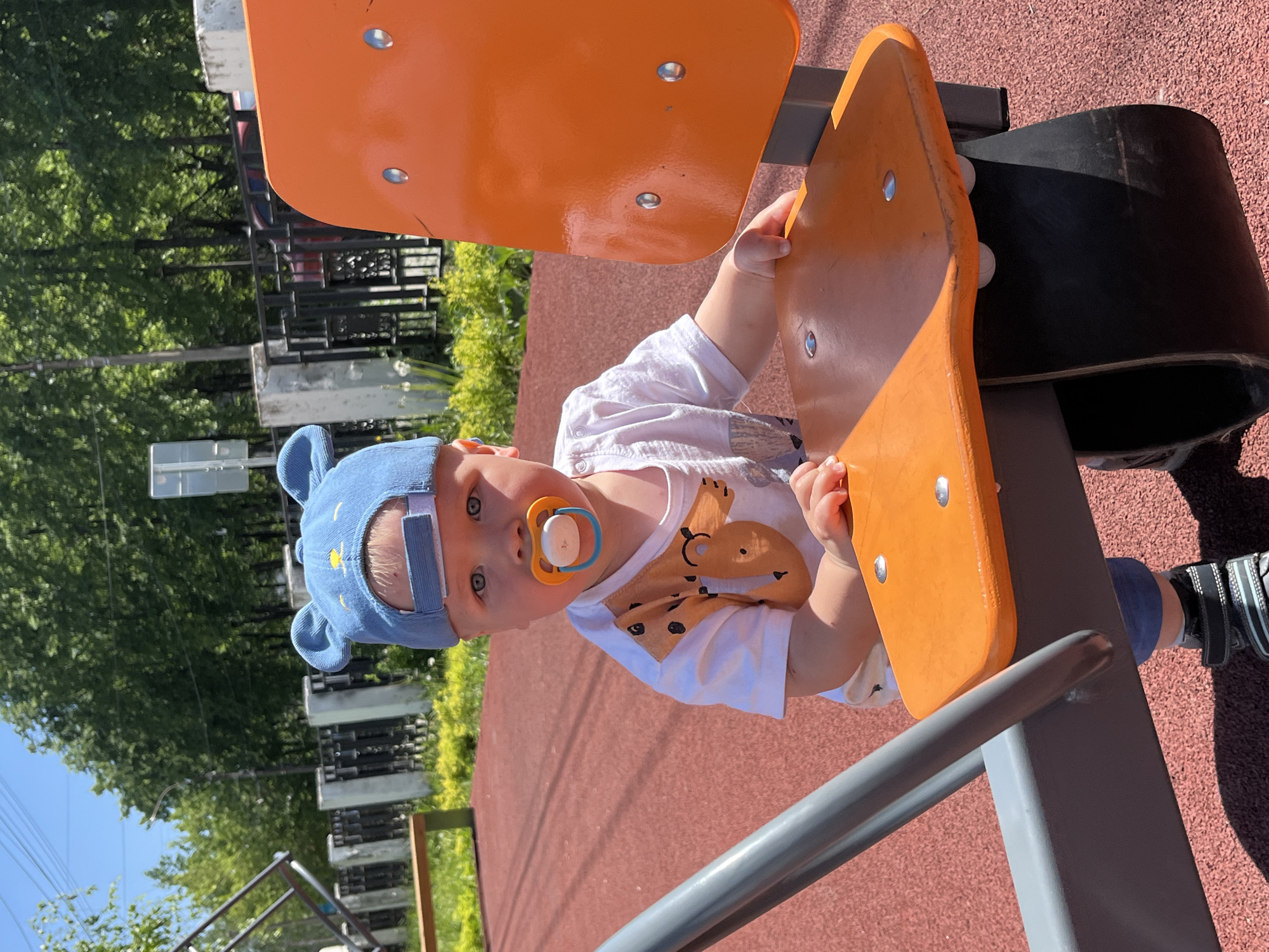 A baby in a blue hat with bear ears is standing at a playground in Chaikovsky, Russia. The baby is wearing a white t-shirt with a brown dinosaur print and has a pacifier in its mouth. The playground has orange and gray equipment on a reddish-brown surface. Trees and a fence are visible in the background.