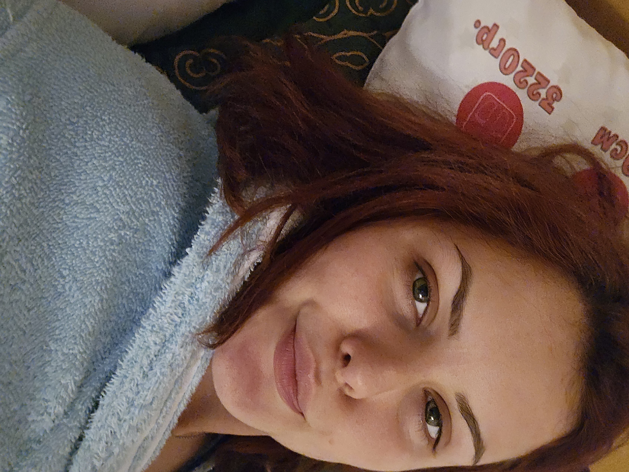 In this close-up, over-the-shoulder shot, a young woman with reddish-brown hair and green eyes is lying down, looking at the camera. She is wearing a light blue terrycloth robe. To her left, a white pillow with orange lettering and a red circular logo is visible. The lettering on the pillow reads "320rp." and "CM." The background consists of dark green fabric with a swirling gold pattern. The image appears to have been taken indoors, possibly in a residential setting. The scene does not provide any specific geographical indicators to determine its location within Golyanovtsi, Bulgaria.