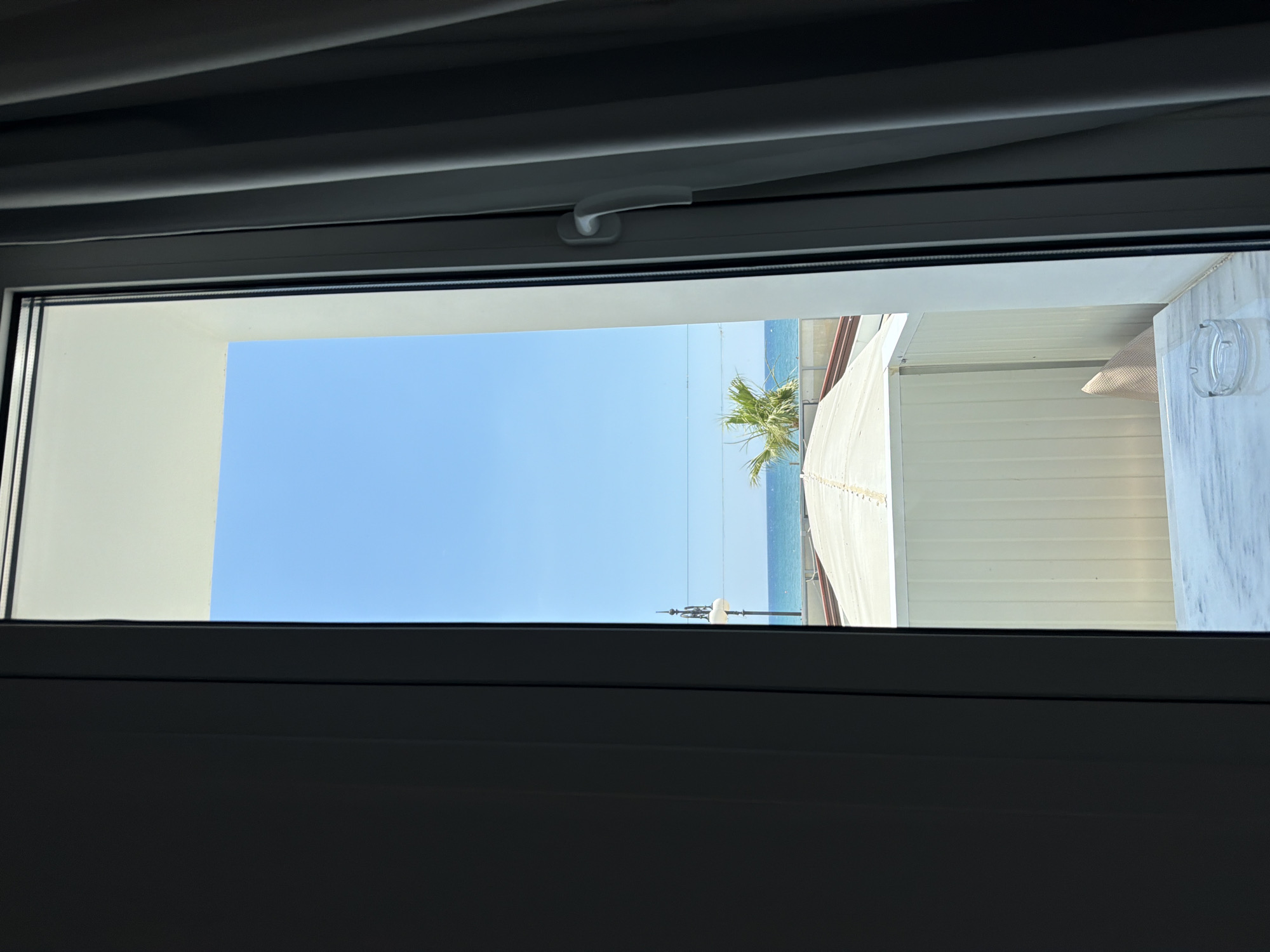 A bright blue sky is visible through an open window. In the distance, the ocean meets the horizon. A palm tree is partially visible on the left side of the image. Below the sky and ocean is a row of white buildings with peaked roofs. A white awning stretches across the top of one of the buildings. The window frame is white, and a gray curtain is visible on the right side of the image. A white marble table is in the foreground, with an empty glass ashtray on it.