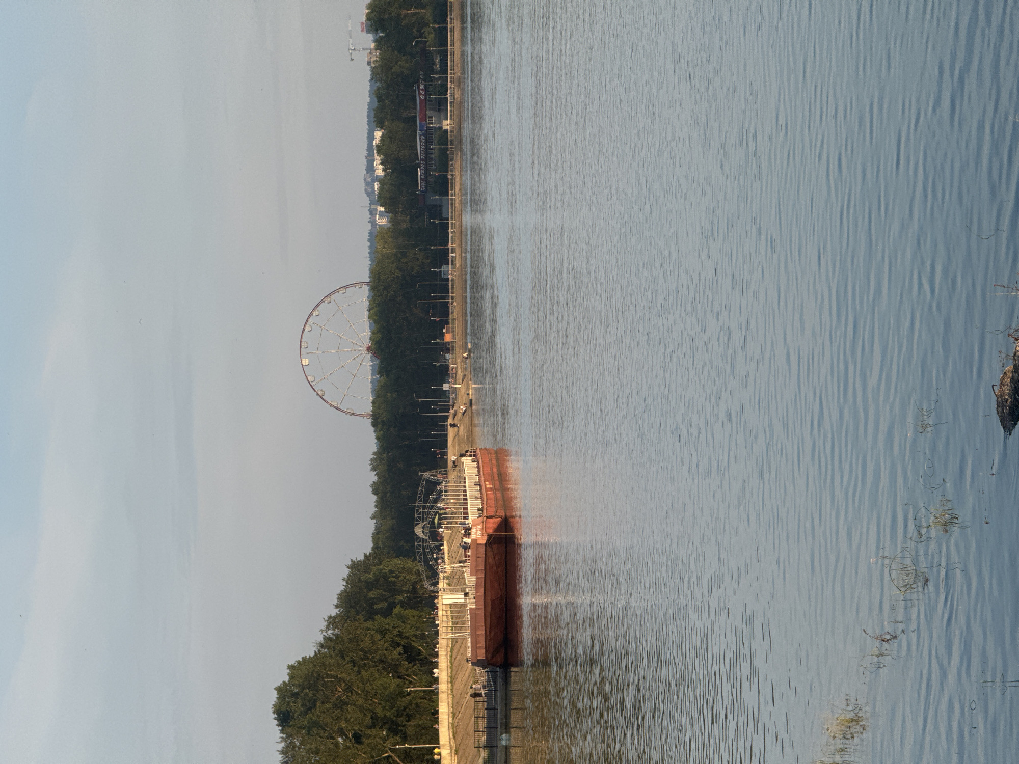 The image captures a serene waterfront scene in Irkutsk, Russia, likely during the late afternoon or early evening. In the foreground, the calm waters of a river or lake reflect the soft light, with small ripples disturbing the surface. On the left bank, a long, flat-topped barge, painted a deep red, is moored to a quay. The barge appears to be a floating structure, possibly a restaurant or entertainment venue, with railings and seating visible along its deck.
Beyond the barge, the riverbank is lined with lush green trees, forming a dense backdrop. A paved promenade runs along the riverfront, where several figures can be seen strolling or sitting. Further in the distance, a large Ferris wheel stands prominently against the pale blue sky, suggesting an amusement park or recreational area. The skyline is dotted with buildings and a construction crane, indicating the presence of a city. The overall atmosphere is tranquil and picturesque, with a sense of calm activity along the water's edge.