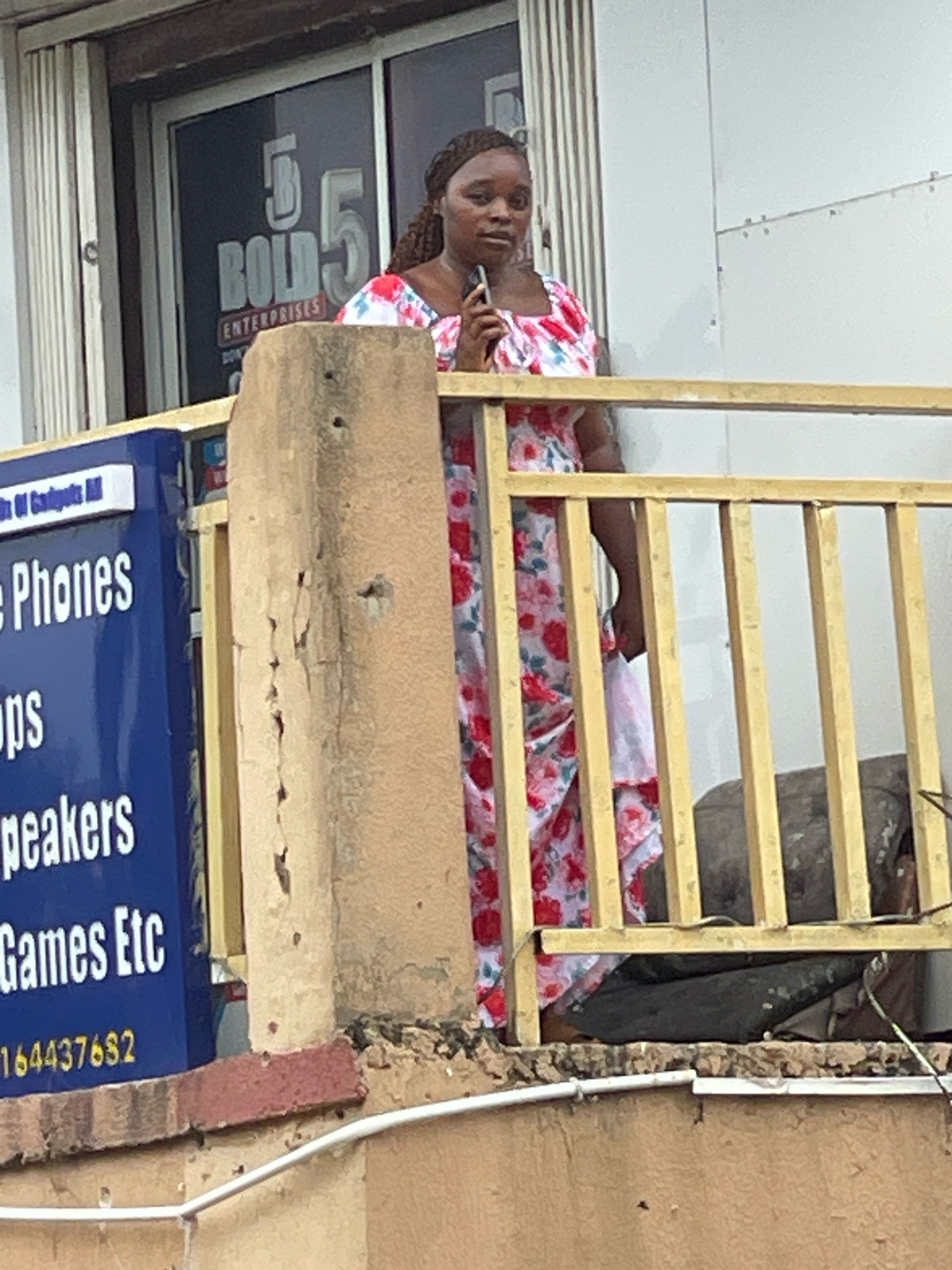 A medium shot captured from below shows a young Black woman with braided hair, wearing a white dress with a red floral pattern, standing on a balcony. She is holding a mobile phone to her ear. Behind her, a window displays signage for "5 BOLD 5 ENTERPRISES" in large gray letters with a red outline. To the left of the woman, a blue sign with white text reads "Phones," "ops," "speakers," "Games Etc," and a phone number "164437682". The balcony railing is made of yellow metal bars. A concrete pillar is partially visible in the foreground. The background appears to be a wall. The scene is likely located in Abuja, Nigeria, given the context of the establishment.