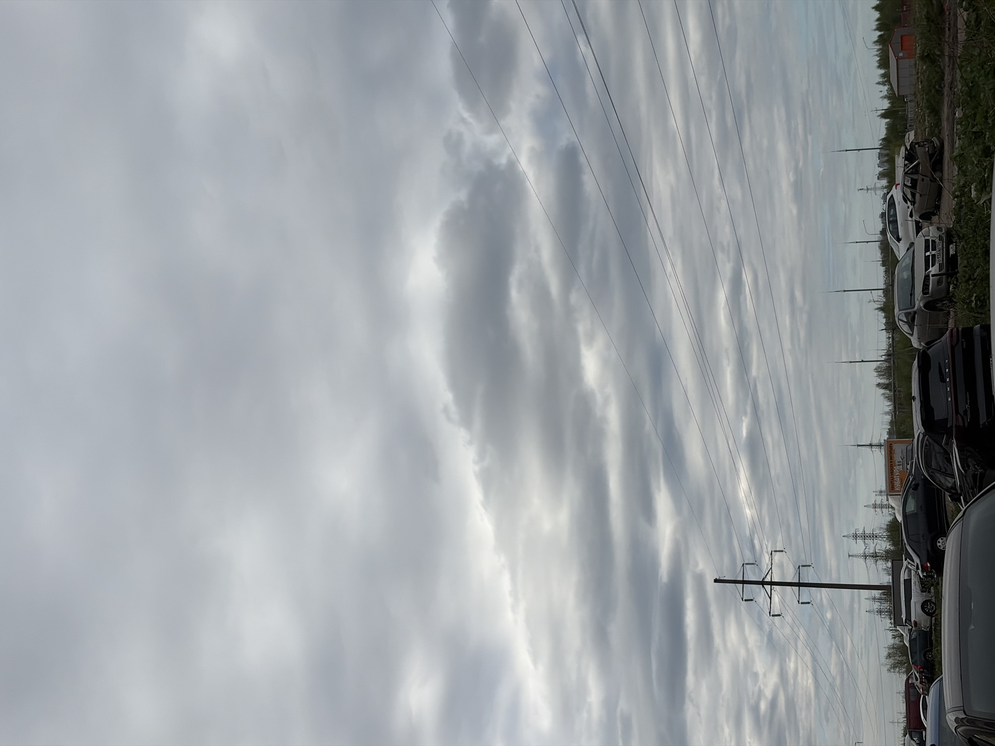 The image depicts a cloudy sky over what appears to be a car parking area or vehicle storage lot. Power lines stretch across the frame. In the background, there are some trees and a few small buildings. The photograph shows an overcast day with the scene captured from a location in Saint Petersburg, Russia.
