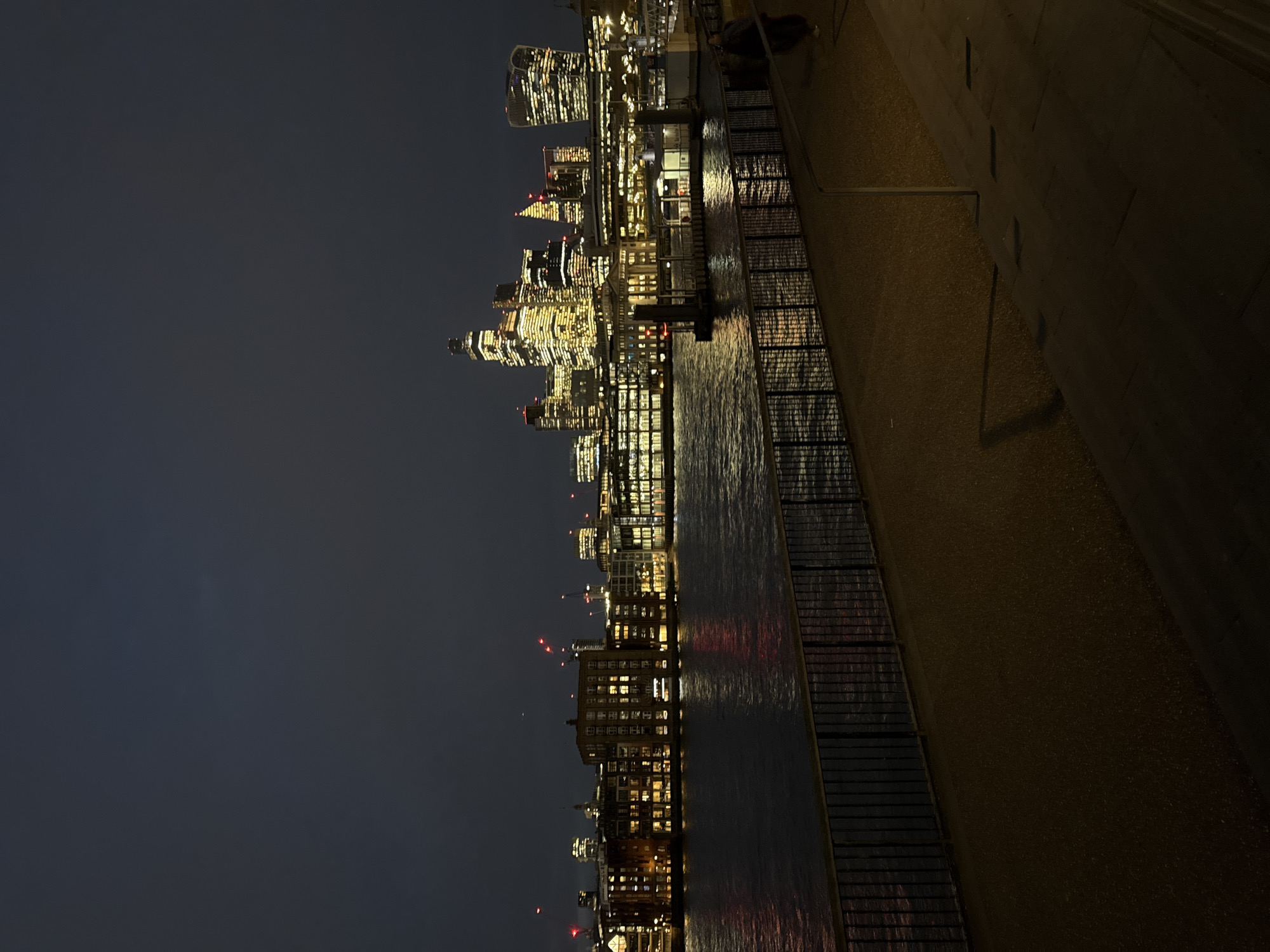 The image displays a nighttime view of the City of London skyline, United Kingdom, observed from a riverside pedestrian walkway on the southern bank of the River Thames. In the foreground, a dark metal railing separates the paved path from the river. The midground consists of the dark, rippled waters of the River Thames, reflecting city lights. A river pier or ferry terminal, including a white structure and dark posts, is visible on the northern bank. The background is dominated by a dense array of illuminated buildings and skyscrapers, forming the distinctive skyline of the City of London. Prominent structures like 22 Bishopsgate, The Gherkin, and The Walkie-Talkie are discernible. Numerous construction cranes with red warning lights are visible above the building tops. The sky is dark and overcast. A single, indistinct individual is partially visible, walking away from the viewer on the far right of the pedestrian path.