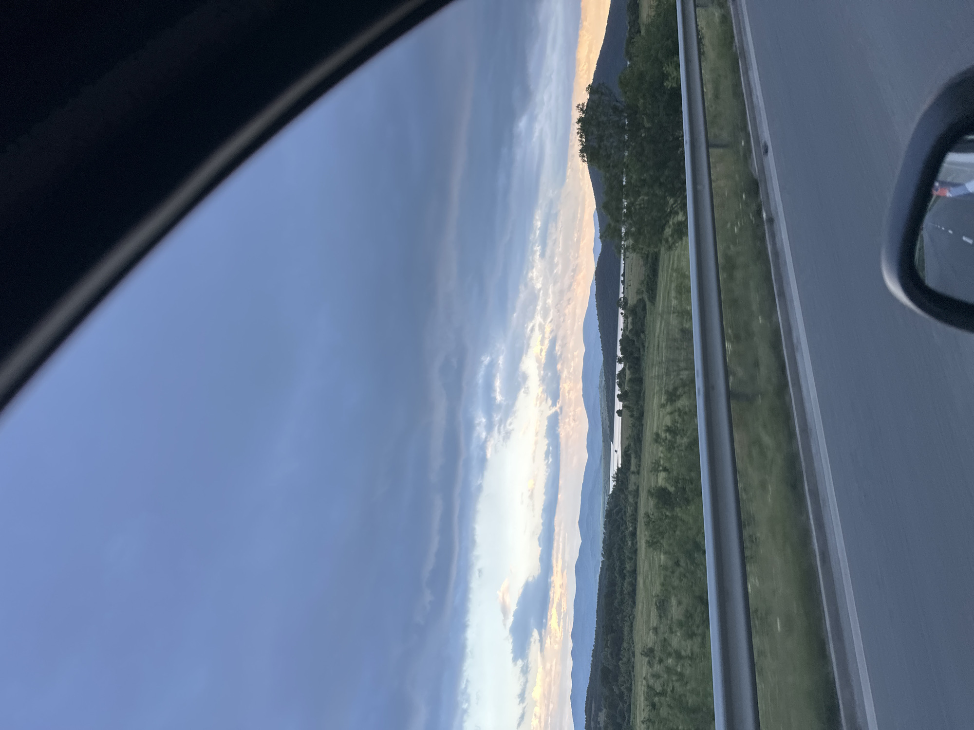 The image shows a view from inside a car on a highway near Konare, Bulgaria. The landscape includes rolling hills, a long lake, and a sky with dark, dramatic clouds. The road has a divider with trees and vegetation in between the lanes. A side-view mirror is visible, reflecting the road behind. The photo captures the scenery from the car window as the vehicle is driving.