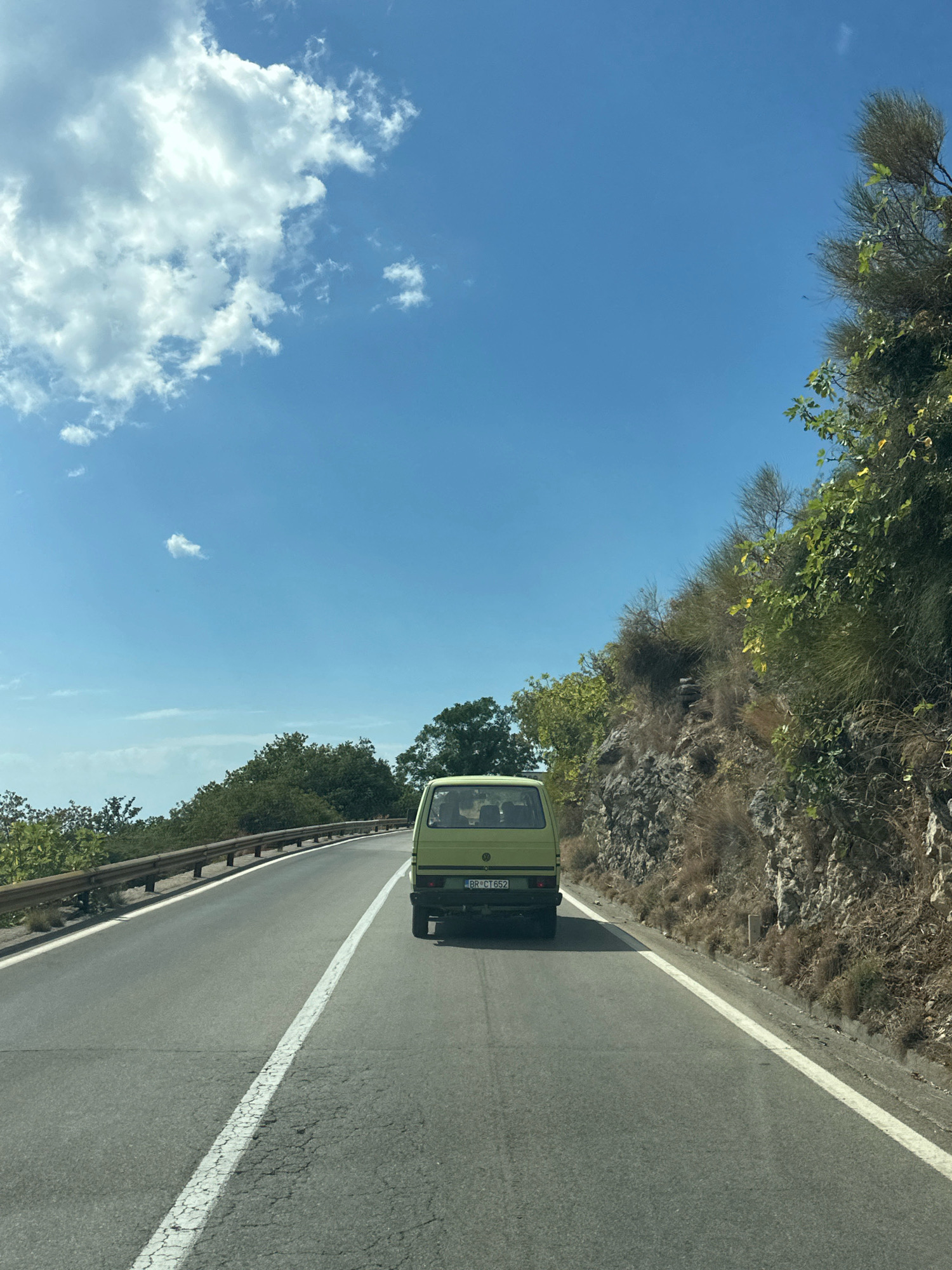 A light green vintage van drives down a paved road in Prijevorac, Montenegro. The van is traveling away from the camera, heading towards a curve in the road. The van has a license plate that reads "BR CT 652". The road is bordered by a guardrail on the left and a rocky, vegetated hillside on the right. The sky is blue with scattered white clouds. Trees and bushes line the left side of the road, and the hillside on the right is covered with dry grass and scrub brush. The overall scene depicts a sunny day on a scenic mountain road.