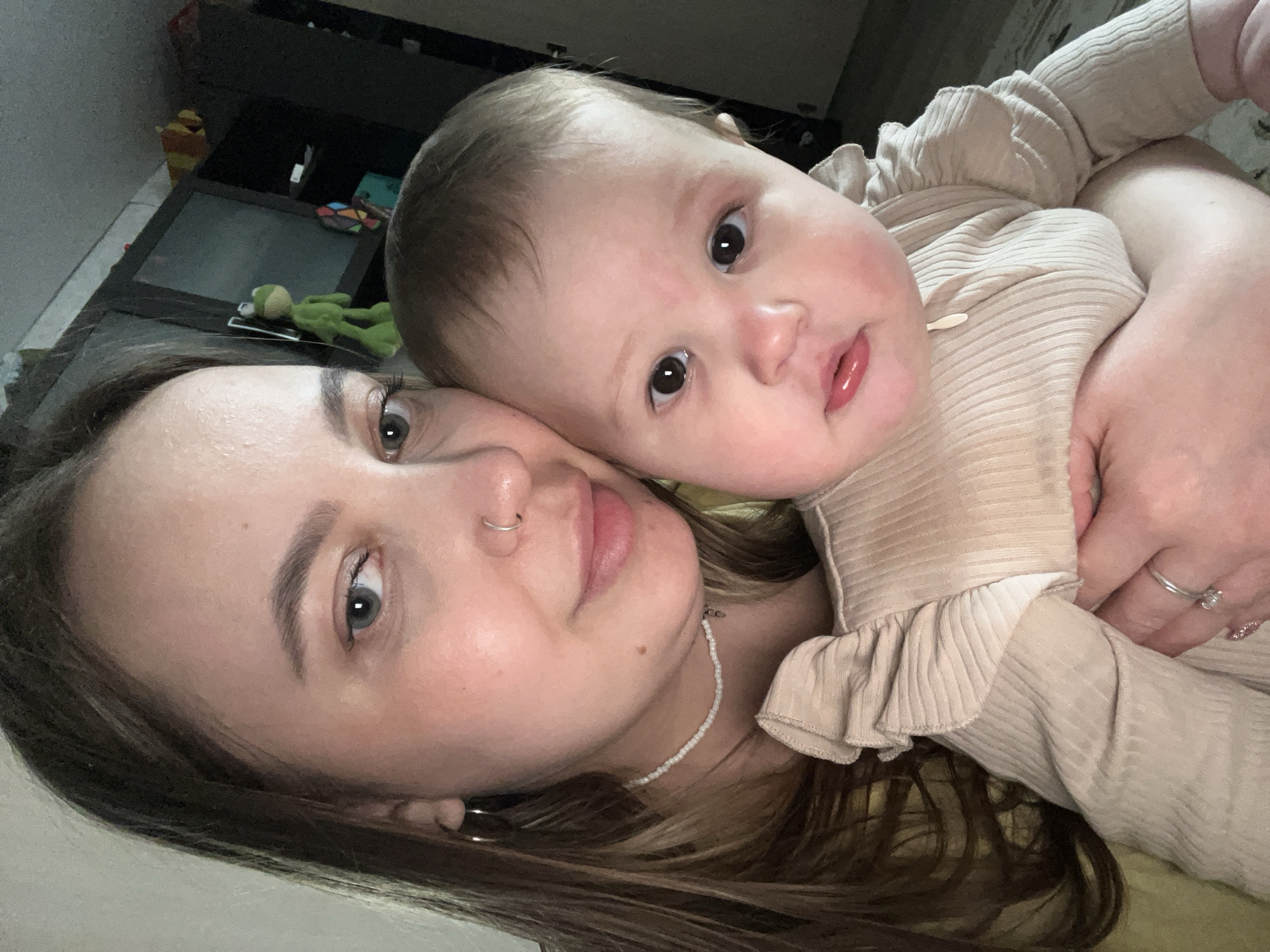 A selfie shows a light-skinned woman with brown hair and blue eyes holding a baby with light skin and brown hair. The woman has a nose ring and is wearing a beaded necklace. The baby is wearing a beige ribbed shirt with ruffles. The background includes a black shelf with toys and a green frog stuffed animal. The location in Novopokrovskaya, Russia, cannot be determined from the image.
