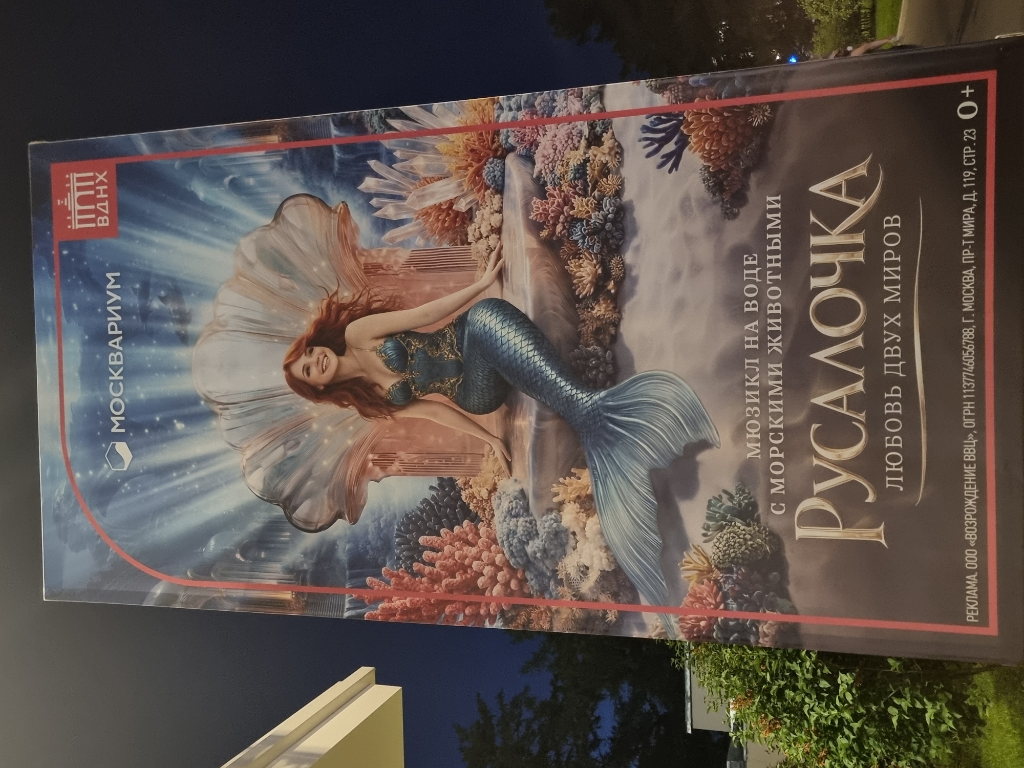 This is a vertical, full-body portrait of a digital painting advertisement for a musical performance titled "Ruslan and Lyudmila: Love of Two Worlds" at the Moscow Aquarium, located at VDNH in Moscow, Russia. The artwork depicts a red-haired mermaid with a blue and gold scaled tail and a matching corset, seated on a rock formation surrounded by colorful coral and crystals. She is looking to her left with a slight smile. The background features a dreamlike underwater scene with a large pearl shell and cascading water. The text on the poster is in Russian and includes the name of the musical, the venue, and advertising details. The poster is illuminated and appears to be a large outdoor advertisement.