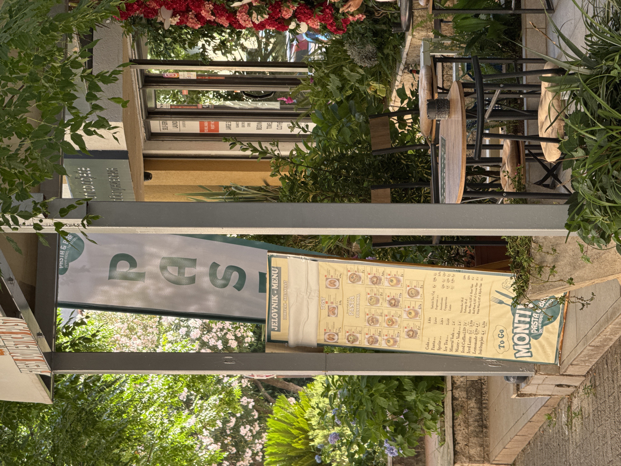 The image shows an outdoor seating area of a restaurant called "MONTI PASTA & BAR" located in Budva, Montenegro. A prominent menu board is displayed on the left, showcasing breakfast items and pasta dishes with prices in Euros. The menu board features the words "JELOVNIK - MENU" and "DORUČAK - BREAKFAST". The restaurant's signage is visible at the top, displaying "FRESH DOMESTIC PASTA".
In the background, the entrance to the establishment is visible with large glass doors. Signs on the door indicate "SELF-SERVICE LAUNDRY" and various services offered. Several tables and chairs are set up for outdoor dining, some with cushions. Lush green plants and flowering bushes surround the seating area, with a decorative floral arrangement of red and pink flowers on the right. The overall atmosphere suggests a relaxed, daytime setting.