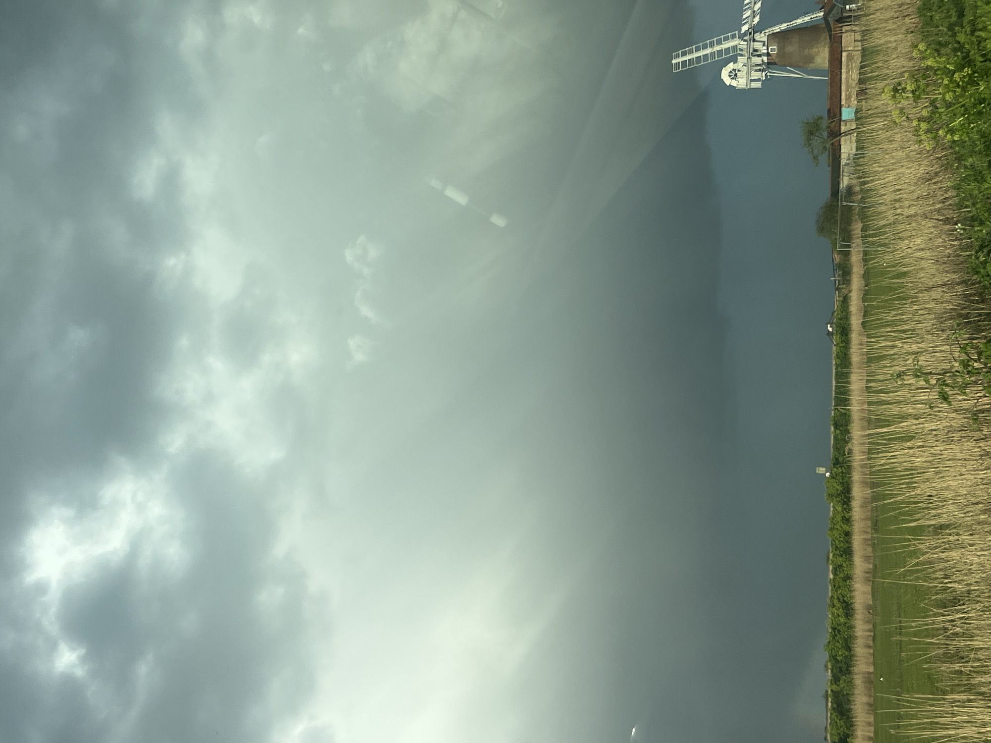 The image depicts a landscape in Broadland, United Kingdom, dominated by a grey, overcast sky and a windmill. The lower right of the frame is occupied by a field with a dirt path running along its edge, bordering a dense line of green vegetation and tall reeds. The windmill stands in the distance, its blades visible against the backdrop of the dark sky. The perspective suggests the photo was taken from a low vantage point, possibly from inside a vehicle looking out, as there are hints of reflections on the sky. The overall scene is rural and muted in color, conveying a sense of a potentially stormy or cloudy day.