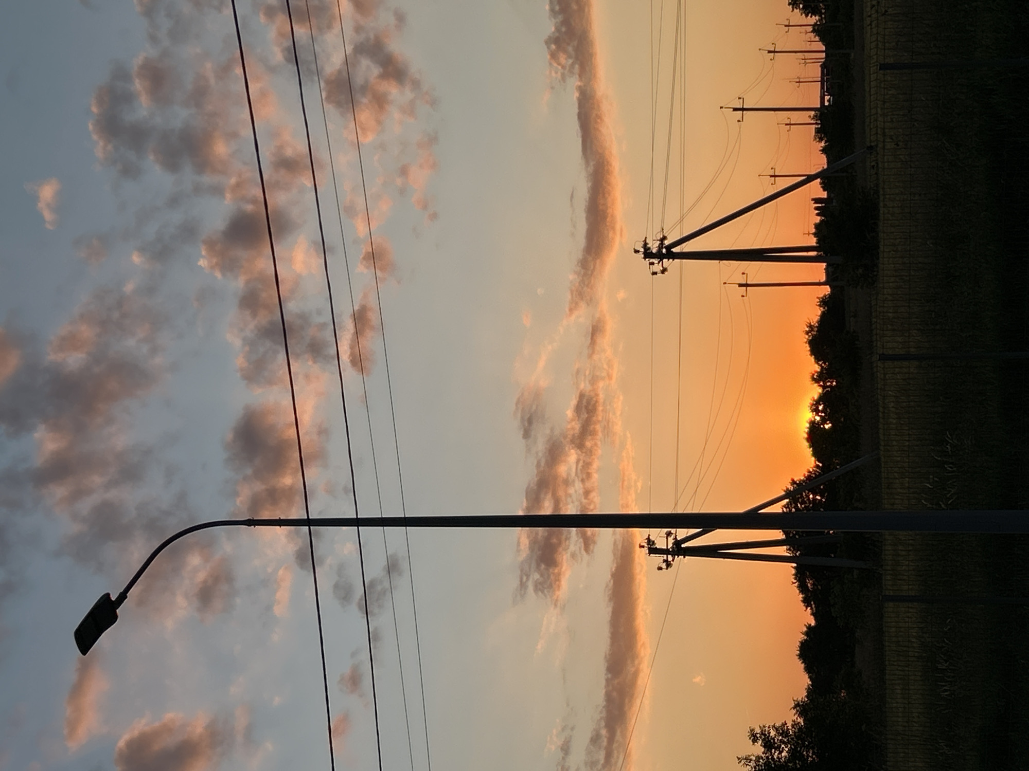 The image captures a sunset in an urban or suburban fringe environment. The sky is a gradient of warm oranges and pale blues, with scattered clouds tinged with pink and orange hues. Dominating the foreground is a tall, dark streetlamp with a curved arm. Several sets of power lines stretch horizontally across the frame, supported by utility poles of varying heights and designs. Some poles are tall and angular, while others are simpler with crossbars. In the middle ground, a line of dark trees forms a silhouette against the vibrant sunset. The ground appears to be covered in grass and vegetation, with a wire fence running horizontally across the lower portion of the image. The overall scene conveys a sense of transition, possibly at the edge of a developed area as day turns to night.