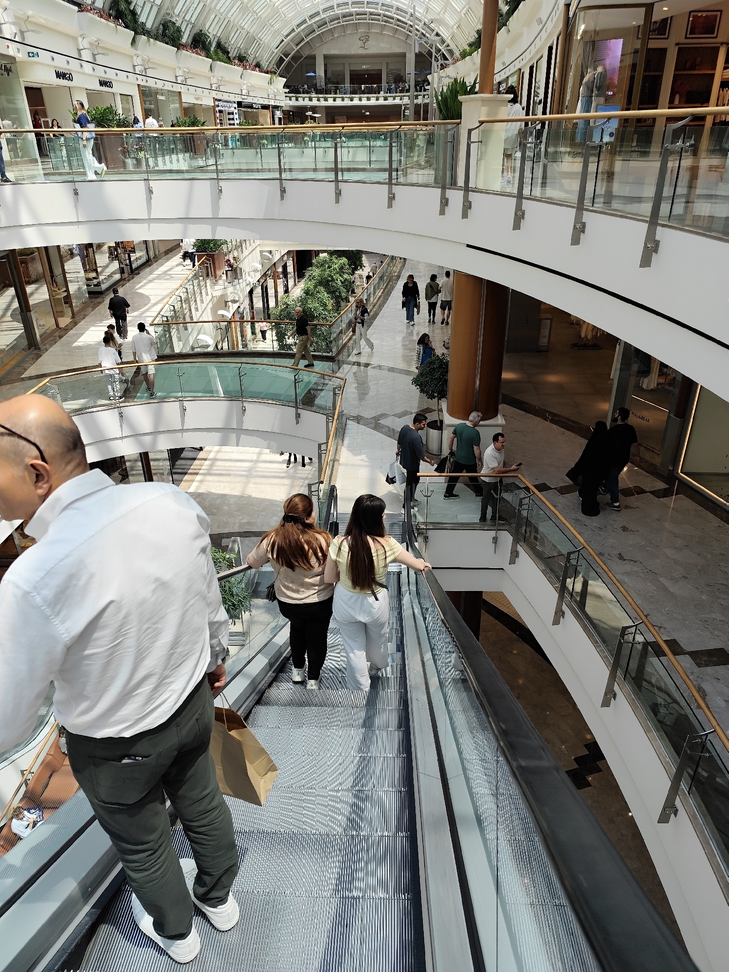 The image captures a multi-level shopping mall interior, possibly located in Sarıyer, Turkey, as indicated by the location data. The mall features a bright, airy environment with a glass-paneled ceiling allowing ample natural light to filter through. The architectural design incorporates multiple levels connected by escalators and walkways with glass railings. Various retail stores are visible along the walkways.
Several people are present within the mall. A man in a white shirt and green pants, carrying a brown paper bag, stands on the escalator. Two women are on the escalator, one wearing a tan shirt and black pants, and the other wearing a light green top and white pants. There are people walking on the various levels. Some are shopping, while others seem to be passing through. A group of people are near a store entrance. The environment includes decorative plants and trees placed strategically throughout the mall, adding to the aesthetic appeal.