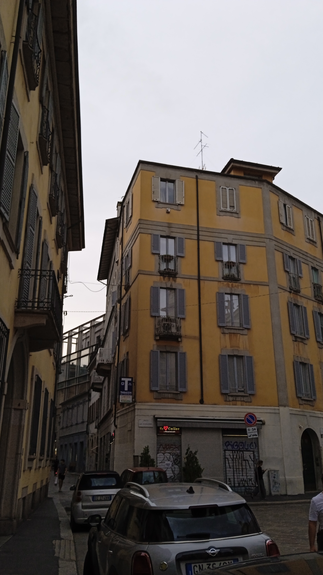 The image depicts a street scene in Milan, Italy, on an overcast day. In the foreground, two silver cars are parked, one of which is a Mini Cooper with a license plate starting with "GN ZC". A pedestrian wearing a white shirt is partially visible on the right, standing near the cars.
The main focus of the image is a tall, multi-story building with a yellow facade. This building is situated on a corner, with another building of similar height and yellow color on the left, creating a narrow street between them. The yellow building features numerous windows with gray shutters, some of which are open, revealing balconies with iron railings. There are also two shops on the ground floor. One shop has a sign that reads "❤️ Coffee" and appears to be a cafe. The other shop, labeled "TABACCHI", is a tobacco shop. Graffiti is visible on the storefronts and walls of the buildings. An antenna is mounted on the roof of the yellow building.
In the background, several other buildings are visible, also exhibiting a similar architectural style. The street is paved with cobblestones. The overall impression is of a dense urban environment.