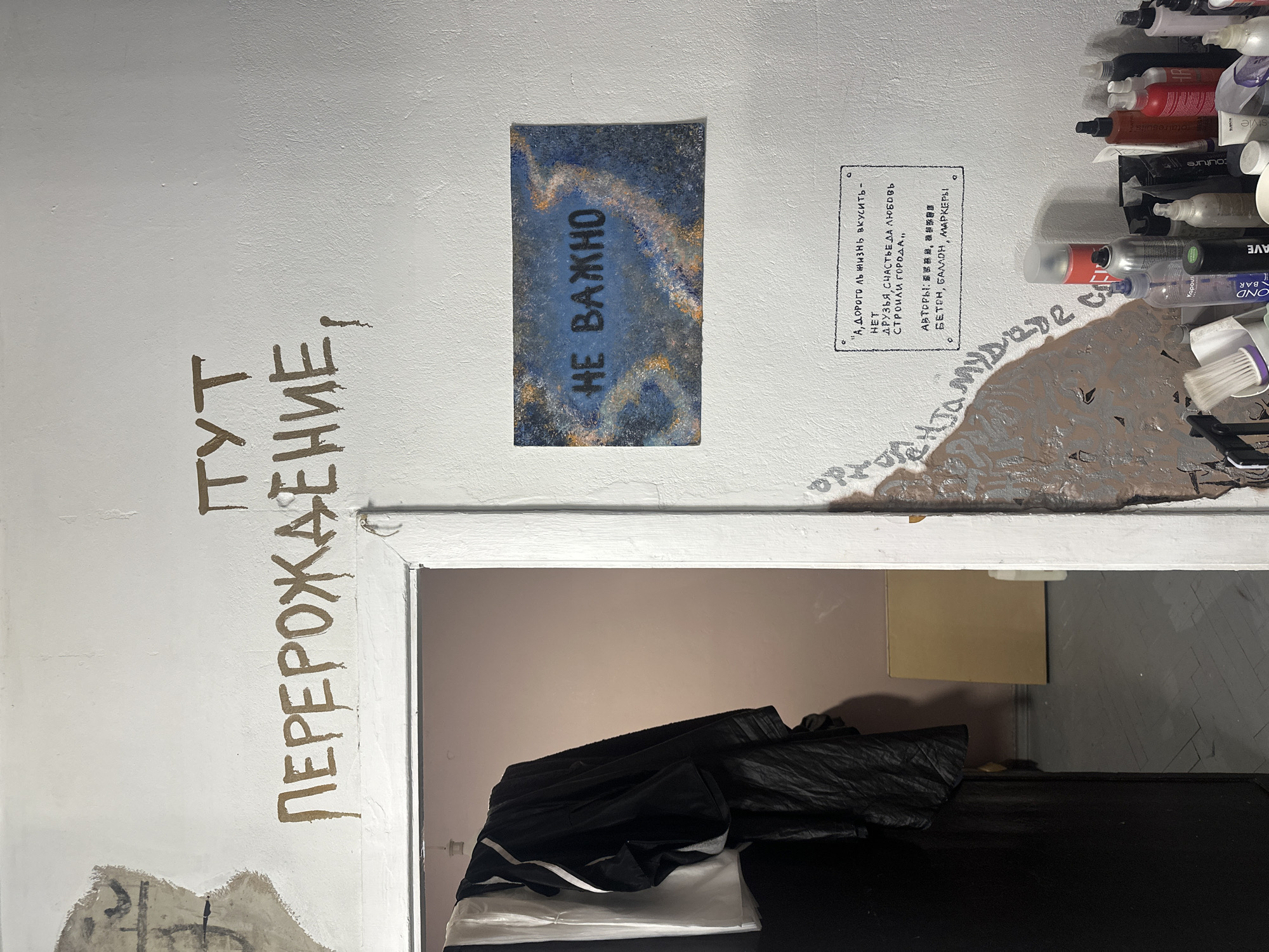 This image captures an interior scene with notable artistic elements on a white wall. The focal point includes the word "ПЕРЕРОЖДЕНИЕ" (Rebirth) painted vertically in gold on the wall near a doorway. Above the doorway, there is artwork: a blue rectangle with "НЕ ВАЖНО" (Not Important) printed on it. To the right of that, a quote inside a box is written in Russian: "А, ДОРОГО ЛЬ ЖИЗНЬ ВКУСИТЬ- НЕТ ДРУЗЬЯ СЧАСТЬЕ ДА ЛЮБОВЬ СТРОИЛИ ГОРОДА," followed by "AUTHORS: Бетон, баллон, маркеры." (Concrete, spray, markers). The right side of the image displays assorted bottles and paint cans cluttered on a ledge. The doorway leads to a room with discarded bags and a glimpse of a dark floor and a light-colored cabinet. The wall also shows signs of disrepair, with patches and paint splatters. Moscow, Russia, is associated with this image due to Cyrillic text.