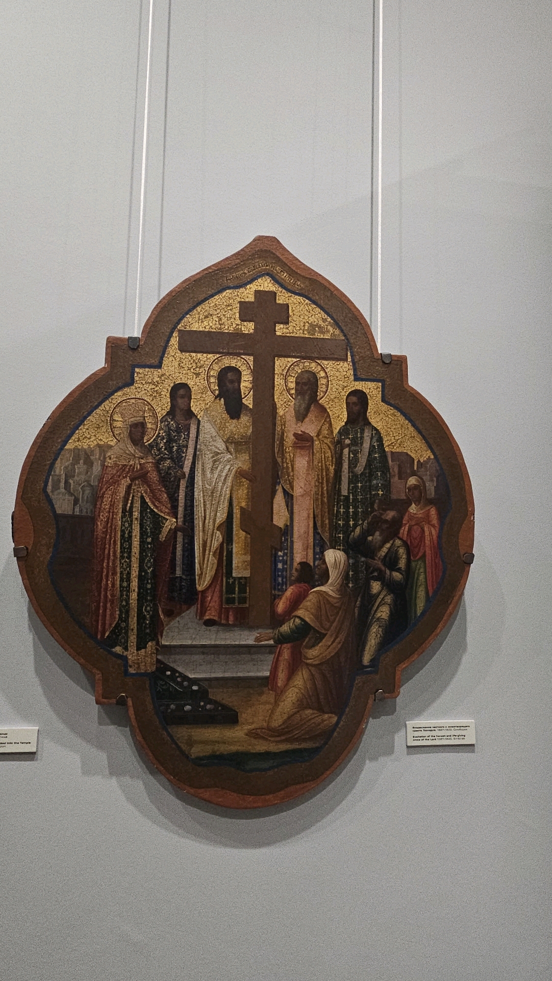 The image shows a religious icon depicting a scene with several figures gathered around a cross. The icon is shaped like a quatrefoil and is framed by a dark wooden border. The central element is a dark brown cross with a golden, mosaic-like background behind it.
Six figures are visible in the icon. On the left, a richly robed figure with a halo stands, holding a scepter or staff. Next to them, another robed figure in a dark patterned garment stands. To the right of the cross, three robed men with halos are positioned. One man, wearing a white and gold robe, has his arm extended towards the cross. Another man, in a pink and gold robe, stands with his hands clasped. A third man, in a green patterned robe, is also present. In the foreground, a woman kneels, her head bowed, wearing a yellow and brown robe. Behind her, another figure in a red robe and a headscarf looks on, along with two smaller figures.
The background of the icon depicts a muted landscape with architectural elements on the left and a textured, golden mosaic pattern behind the figures. The icon itself is suspended by two thin white wires against a plain, light gray wall.
Below the icon, on the left, a small white label is partially visible with some text in Cyrillic. On the right, another small white label is clearly visible with text in both Cyrillic and English. The English text reads: " icon "Presentation of the Lord in the Temple" 1887-1900. Smirdov. Artist of the Lord 1887-1900. Smirdov."
This artwork is on display in the Ulyanovsk Regional Art Museum in Ulyanovsk, Russia. The museum is known for its collection of Russian and Western European art.