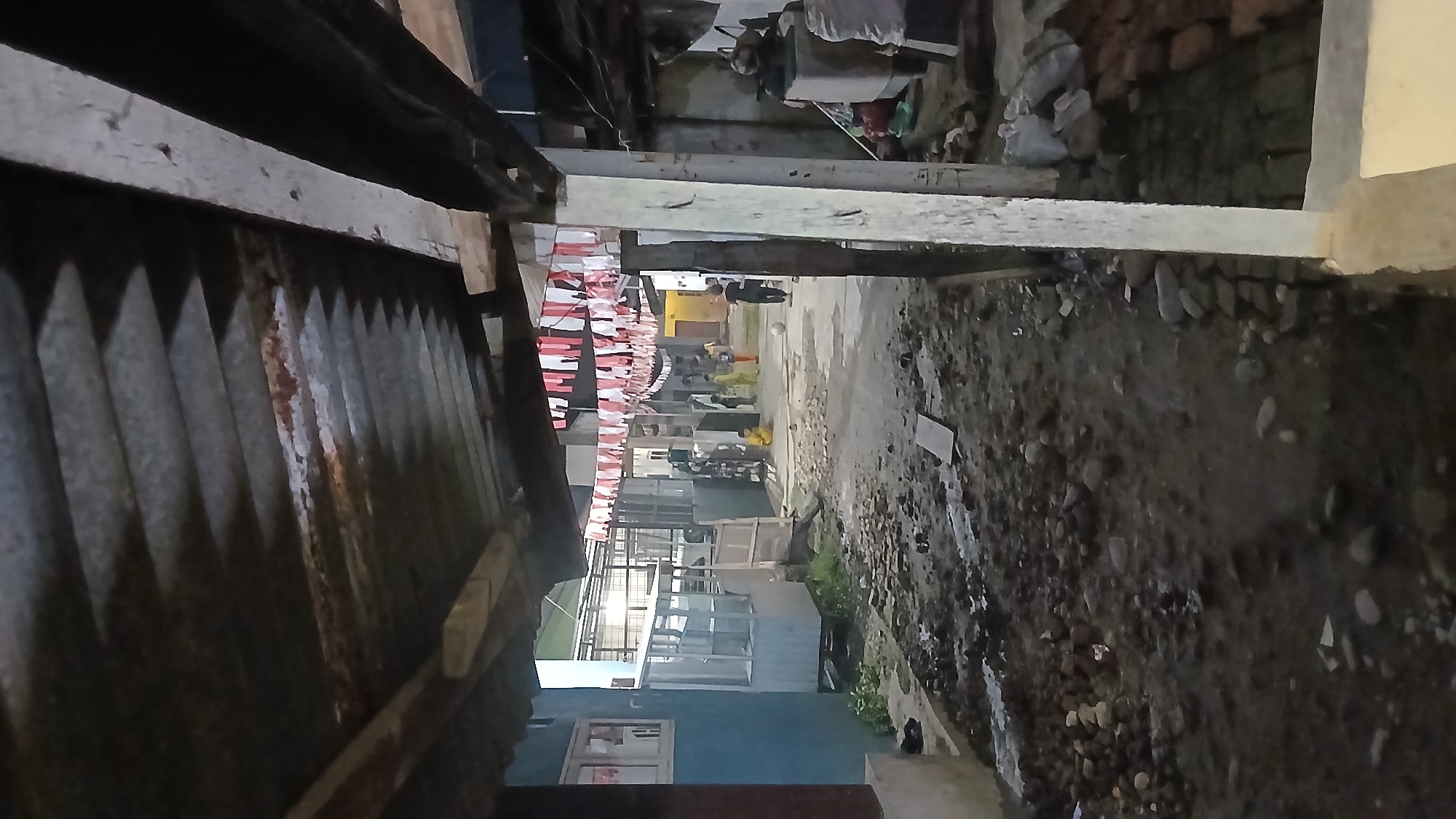 The image depicts a narrow alleyway in Klambir Lima Kebun, Indonesia, characterized by a rough, uneven ground surface composed of dirt and stones. The scene is framed by an overhanging corrugated metal roof, supported by wooden beams. Red and white fabric banners are strung across the alleyway above, suggesting a festive or commemorative occasion. In the background, several children are visible, some appearing to be playing. On the right side of the alley, a washing machine and stacks of materials, including what appears to be sacks and bricks, are present, indicating a residential or working area. The overall atmosphere is that of a utilitarian, possibly impoverished, urban or semi-urban environment.