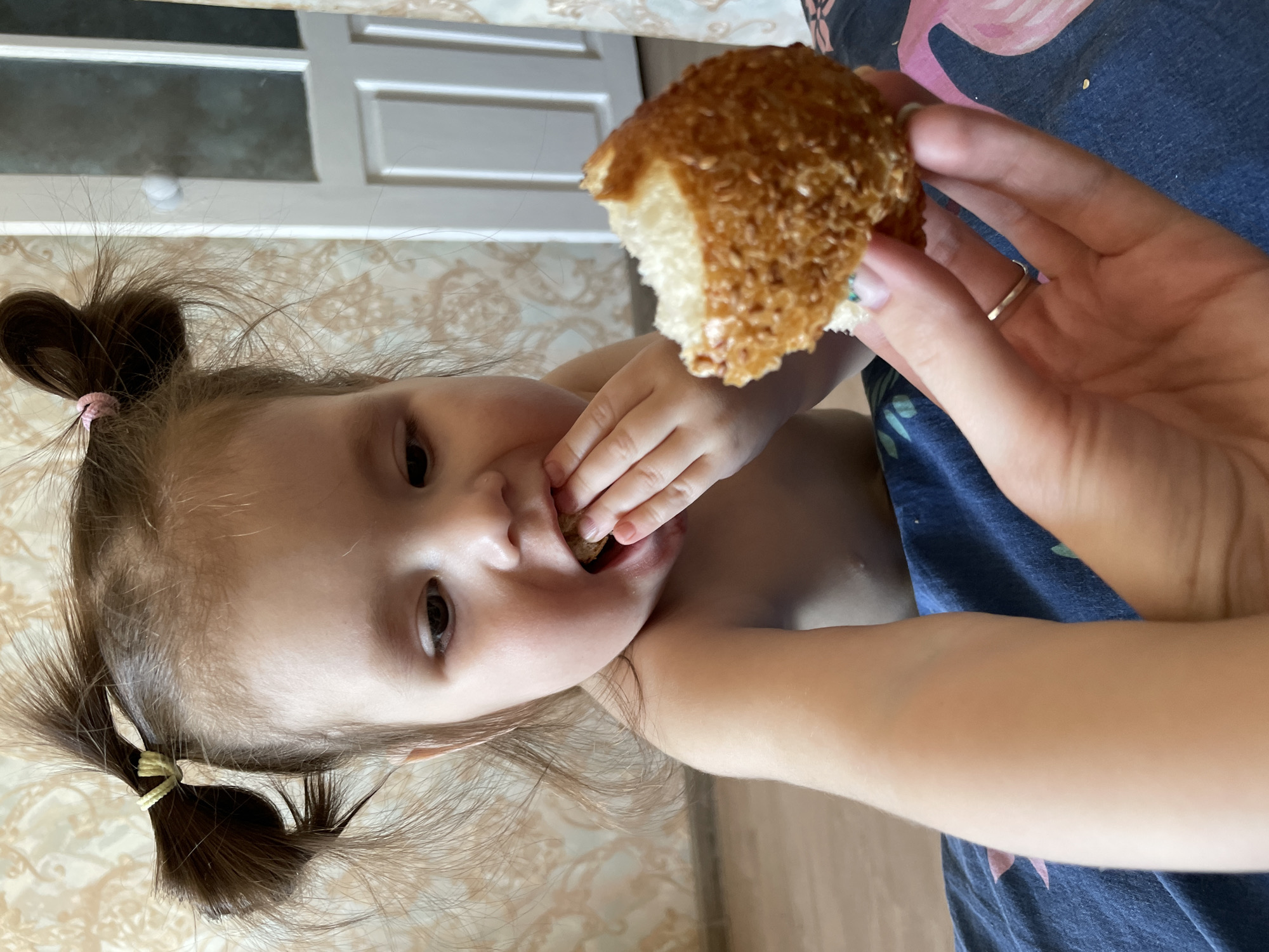 A young child with brown hair tied in two pigtails is eating a piece of bread. The child is positioned in the lower left of the frame, with their face and upper body visible. A hand holding the piece of bread is in the upper right of the frame. The background includes a wall with patterned wallpaper and a white door. A blue sheet with a pink flamingo design is also visible. The location and time are not discernible from the image. The city of Izhevsk, Russia, is not discernible from the image.