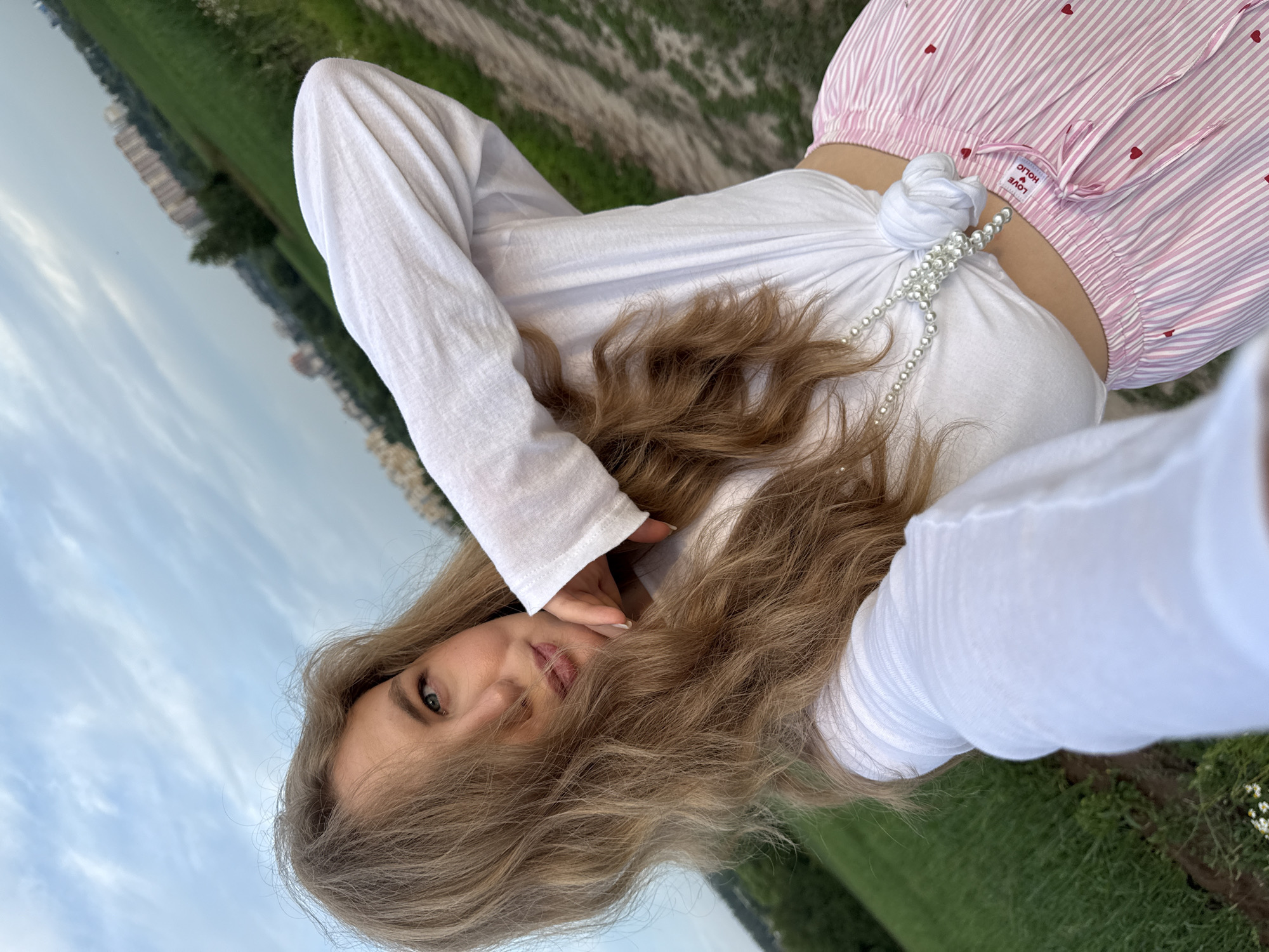 A young woman with long, wavy blonde hair is taking a selfie outdoors. She is wearing a white long-sleeved shirt tied at the waist and pink and white striped pants with small red hearts. A pearl necklace is draped around her neck and over the knotted shirt. Her left arm is raised, with her hand near her face, and her right hand is holding the phone to take the picture. The background consists of a field of green grass, a dirt path, and a cloudy sky with some buildings in the distance. The location is identified as the city of Сновицы, Russia.