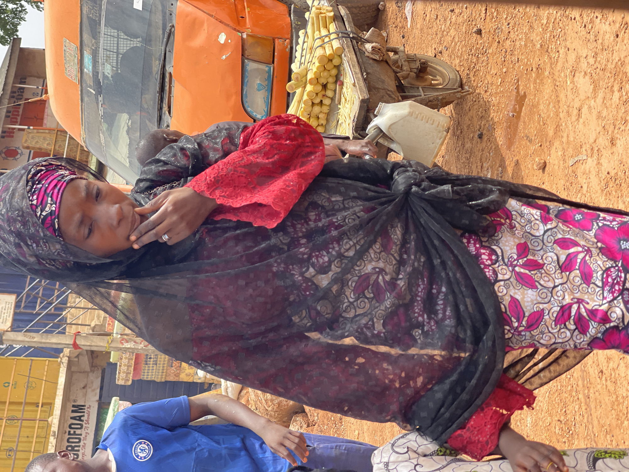 In Zaria, Nigeria, a woman wearing a patterned head covering and draped in a sheer black scarf holds a baby. She is dressed in a floral-patterned top and a red lace-trimmed garment. She has a ring on her finger and appears to be touching her face. Next to her is a cart loaded with sugarcane, attached to a small orange vehicle, possibly a tricycle. The ground is reddish-brown, suggesting soil or unpaved terrain. Parts of buildings and a sign that reads "ROFOAM" are visible in the background. Another person wearing a blue Chelsea FC shirt is partially visible in the lower left corner.