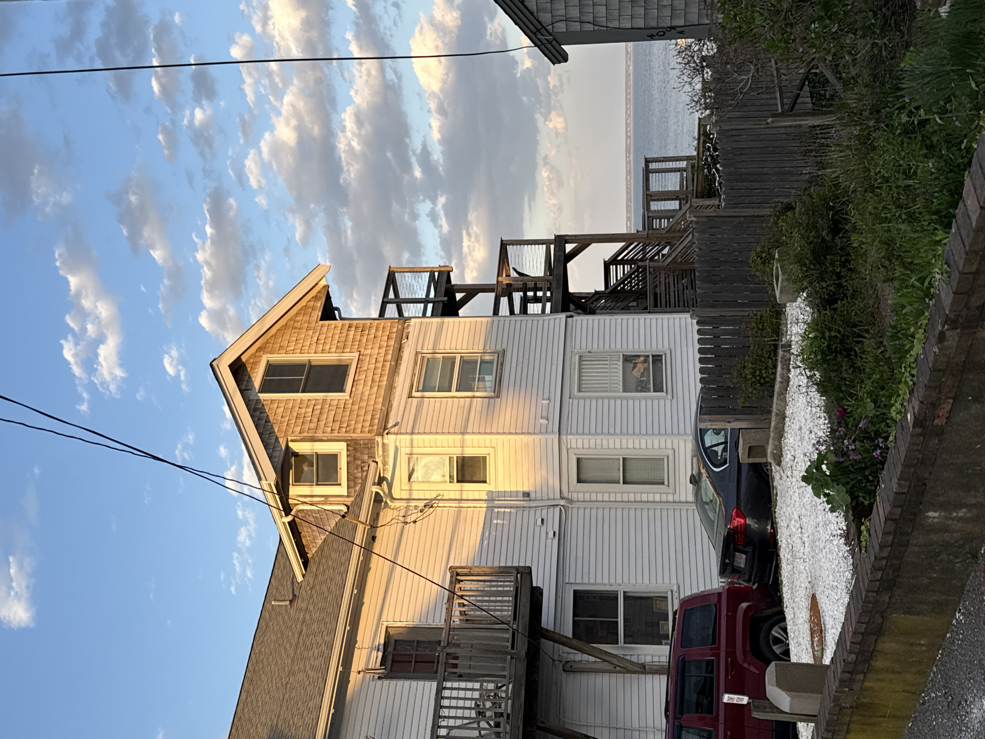 The image shows a three-story house in Provincetown, Massachusetts. The house has a shingled top floor and two white-sided lower floors. There are multiple windows on each floor, and a small balcony on the first floor. A series of wooden stairs and platforms lead to the roof. There are cars parked in front of the house, a red Jeep and a dark sedan. A white gravel path leads to the cars. In the background, the ocean is visible. The sky is blue with scattered clouds. Power lines run across the upper left of the frame. A wooden fence and some greenery are on the right side of the frame. The photo appears to have been taken from a slightly low angle, looking up at the house.