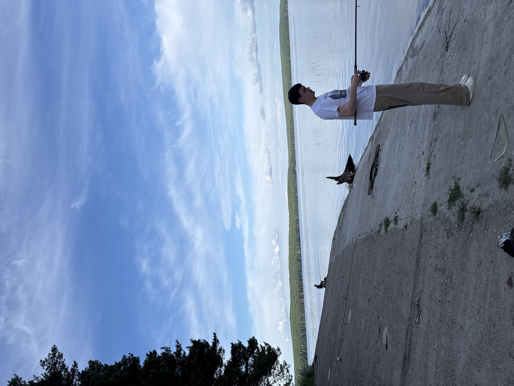 The image, rotated 90 degrees clockwise, captures a young man fishing from a concrete embankment next to a large body of water under a bright, partly cloudy sky in Chaikovsky, Russia.
**Subjects:** The central subject is a young man, likely in his late teens or early twenties, standing in profile. He has short, dark hair and is dressed in a white short-sleeved T-shirt with a dark graphic print on the front, paired with khaki-colored pants and white sneakers. He is holding a fishing rod with both hands, its tip extending over the water. The background features a wide body of calm water, possibly a lake or a broad river, which meets a distant treeline on the horizon. The immediate foreground is a wide, textured concrete embankment, with some small pieces of wood or debris and sparse green vegetation growing along its edges.
**Activity or event taking place:** The man is actively fishing, holding his rod steady and gazing out over the water, suggesting a moment of quiet contemplation or anticipation.
**Notable details:** The scene is illuminated by bright daylight, indicating either late morning or afternoon. The sky is a clear blue, adorned with soft, wispy cirrus clouds and some more defined cumulus clouds, suggesting fair weather. The concrete embankment provides a stable platform for fishing. The distant treeline hints at a natural, expansive landscape beyond the water.
**Visible text:** A graphic print is visible on the front of the man's white T-shirt. The text or specific details of the design are too indistinct to be read or identified clearly.