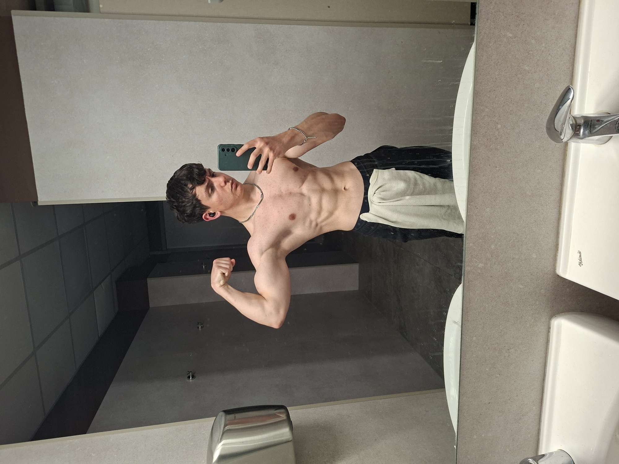A shirtless young man is taking a selfie in a bathroom mirror. He has short dark hair, a necklace, and a bracelet. He's wearing dark pants and a light-colored towel is draped over his side. He's flexing his left arm. The bathroom has a light gray tiled wall and ceiling. A sink with a chrome faucet is visible on the right side of the frame. There is a stainless steel hand dryer in the lower left corner. The location is not identifiable as being in Plovdiv, Bulgaria, based on the visual information provided.