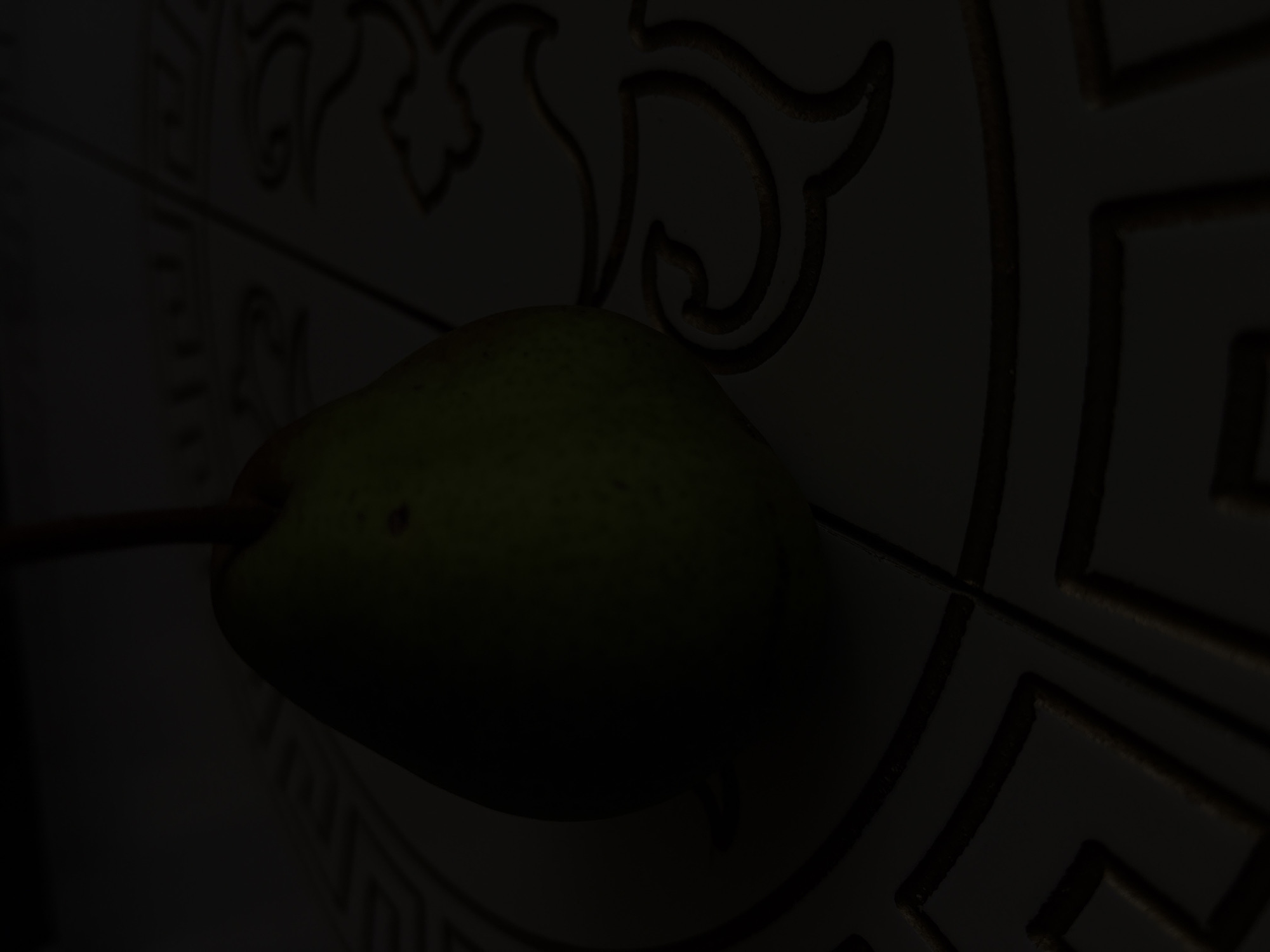 A dimly lit, close-up, overhead shot features a single, green pear positioned on a decorative tiled surface. The pear is the primary subject, with its rounded body and stem visible. The background consists of patterned tiles with raised, golden-colored decorative elements. The lighting is low, casting shadows and obscuring fine details of the pattern. The image appears to be an indoor still life, with no indication of a specific location in Georgiyevsk, Russia, or any people present.