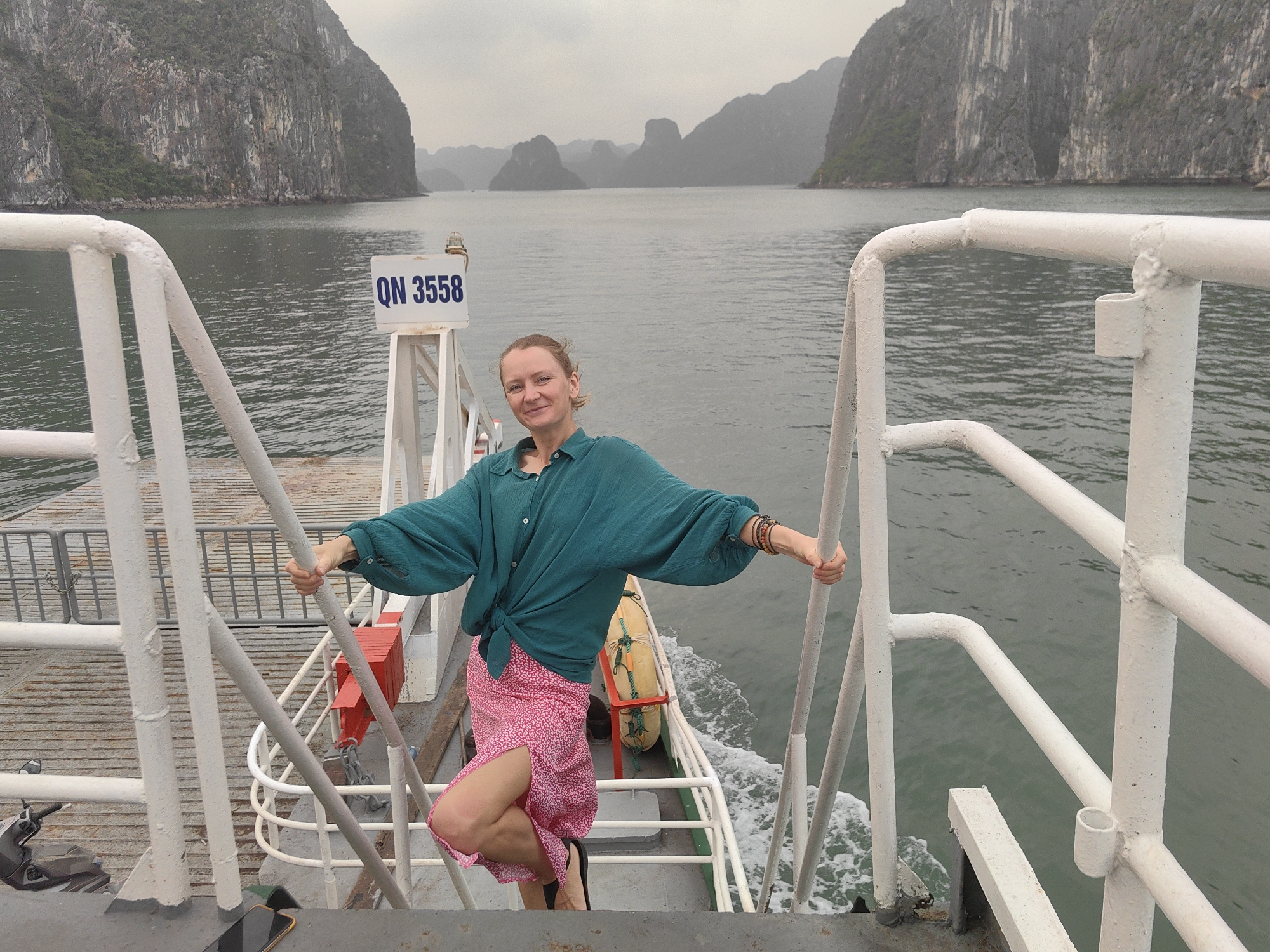 A woman is standing on the deck of a boat in Bai Chay Ward, Vietnam. The boat is surrounded by water and limestone karsts. The woman is wearing a green shirt and a pink skirt. She is holding onto the white metal railing of the boat. A sign on the boat reads "QN 3558". The sky is overcast.