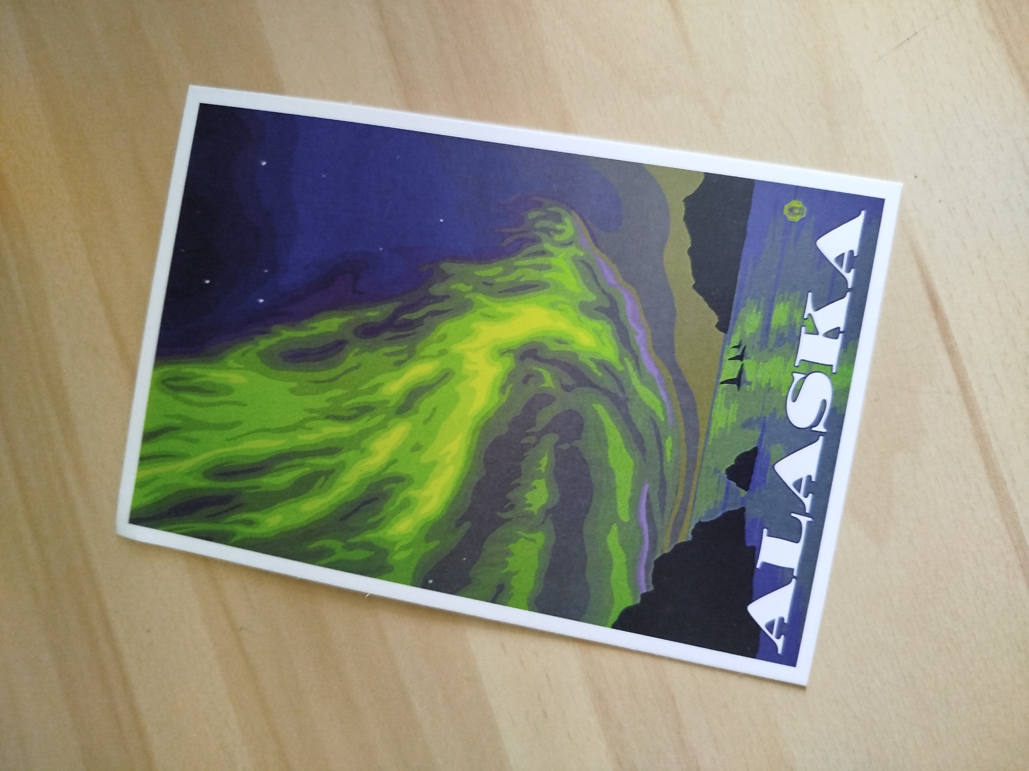 The image is a postcard featuring an illustration of the Aurora Borealis over Alaska. The sky is dark blue with a few stars. The aurora is depicted with shades of green and yellow, flowing across the sky. Silhouetted mountains and bodies of water are also visible. The word "ALASKA" is printed vertically on the right side of the postcard in white letters. The postcard is resting on a light-colored wooden surface.