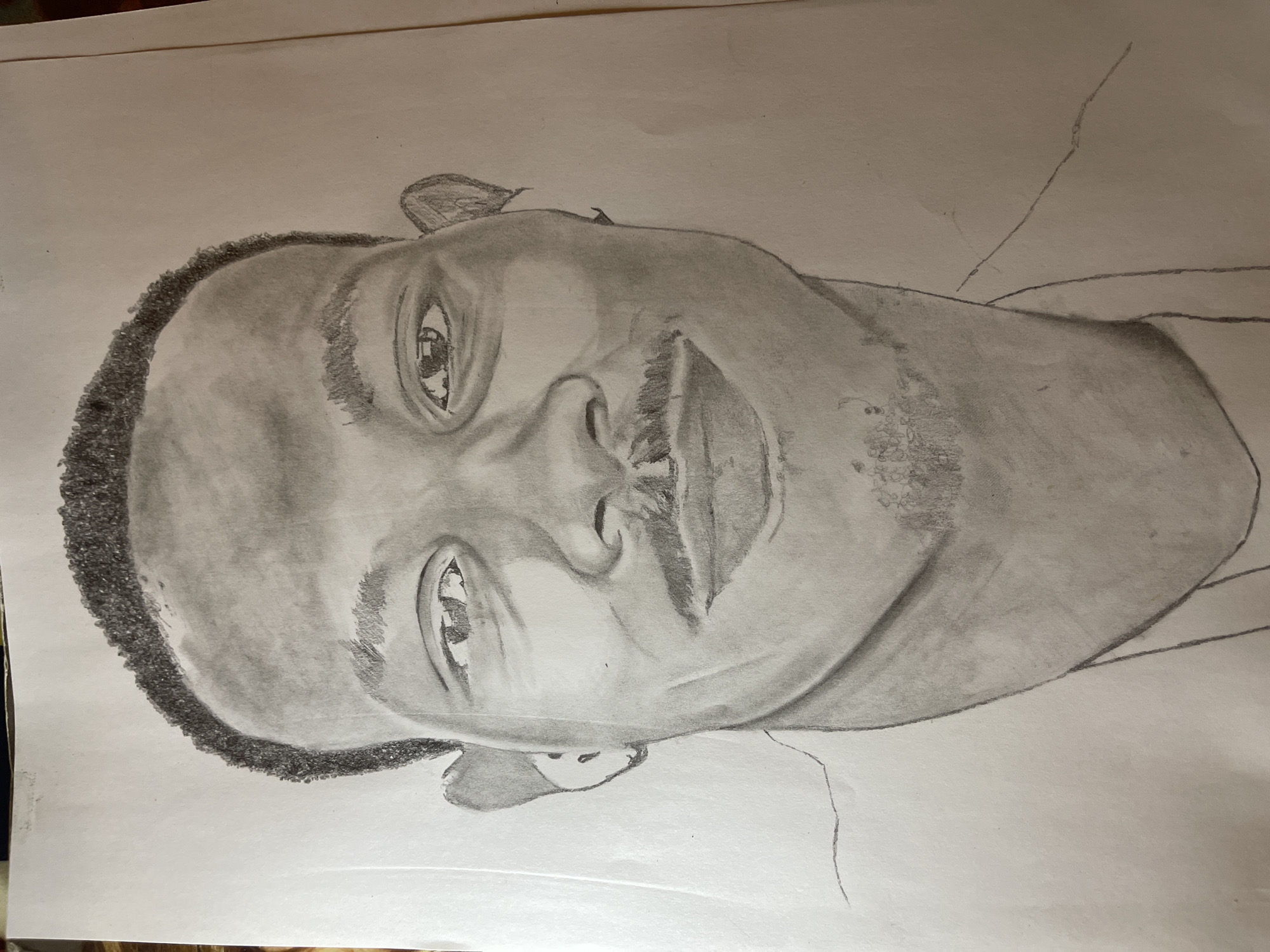 The image is a pencil drawing of a man. The subject is facing the viewer with their head tilted slightly. The drawing features details such as the hair texture, facial features including eyes, nose, mouth, and stubble, and shading to suggest form and depth. The drawing is on a piece of paper, and there are some faint lines indicating an outline of the shoulders. The background is plain white. The location of Jos, Nigeria, is not determinable based on the information in the image.