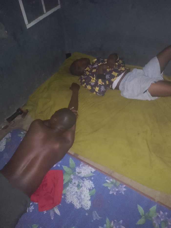 Two people lie on adjacent mattresses. One person is shirtless, lying face down on a floral-patterned mattress, with one arm raised. A red cloth is nearby. The second person, wearing shorts and a patterned shirt, lies face up on a yellow mattress. A flashlight sits near the edge of the mattress. The room is poorly lit with a window visible in the background.
