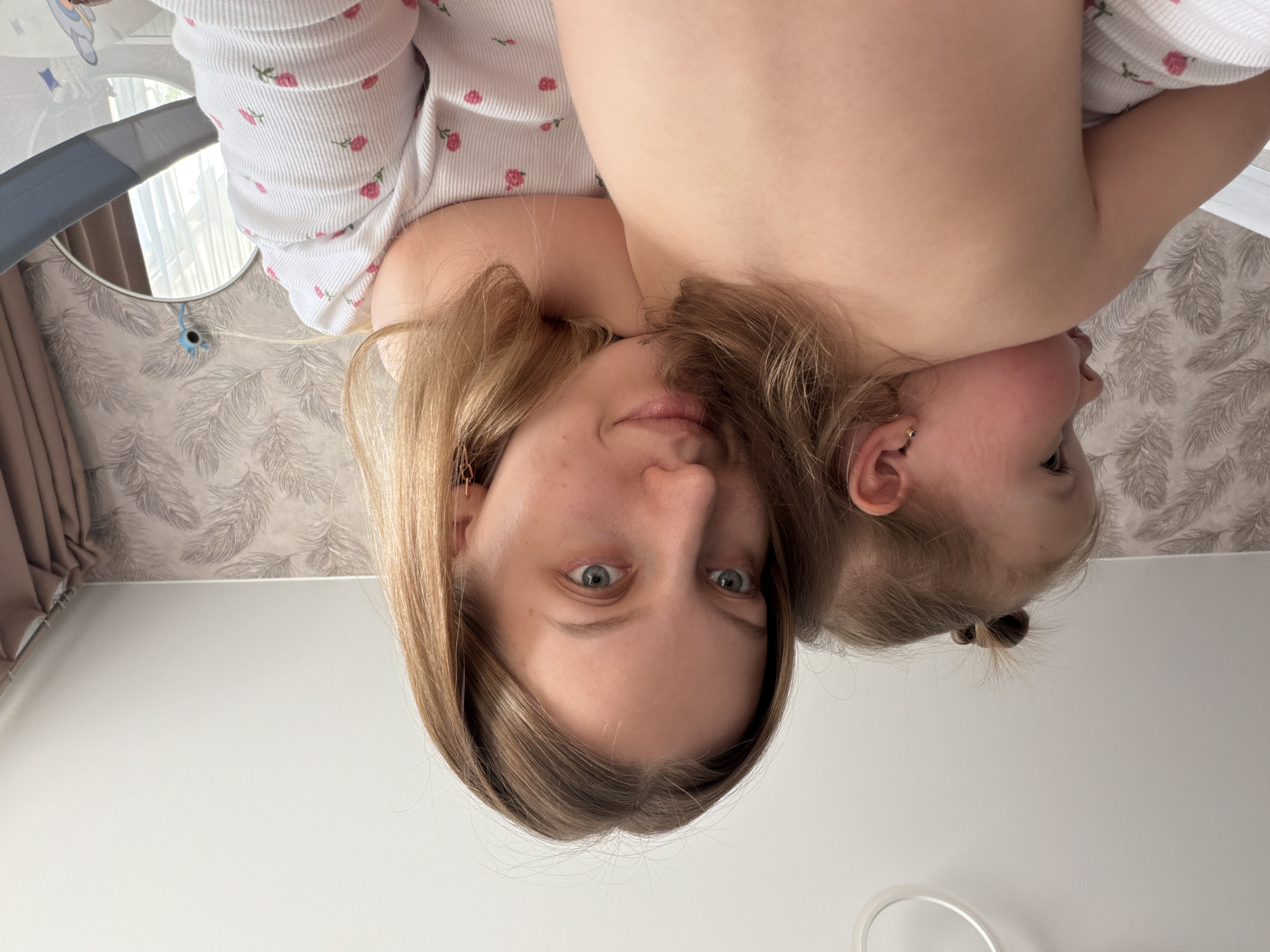 A fair-skinned woman with light brown hair and blue eyes is looking directly at the camera. She is wearing a white top with pink floral patterns. A fair-skinned baby is positioned to her right, with its face turned away. The baby has light brown hair pulled into a small bun. The background includes a wall with a leaf pattern, a grey baby bouncer, and a white wall. The location is Reshetnikovo, Russia.