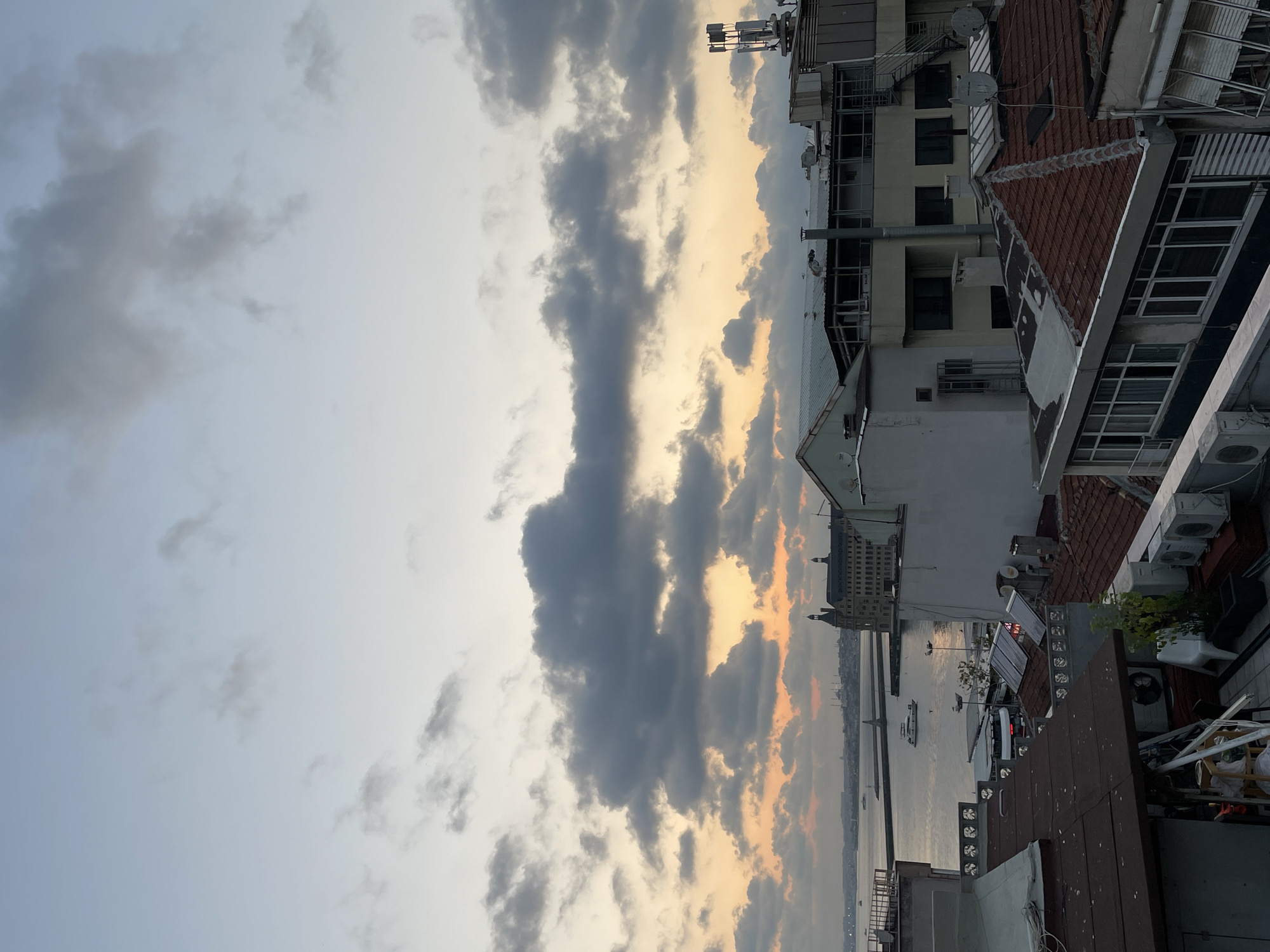 This is a rooftop view of Kadıköy, Turkey, at sunset. The sky is filled with dramatic clouds, tinged with orange and yellow from the setting sun. In the distance, the waters of the Bosphorus are visible, with several boats and ferries traversing the waterway. Across the water, the iconic Haydarpaşa Train Station building is prominent, along with other city structures. The foreground is dominated by the rooftops of various buildings in Kadıköy, showcasing a mix of architectural styles and rooftop clutter, including satellite dishes, air conditioning units, and ventilation pipes. The scene captures a moment of urban tranquility as the day draws to a close.