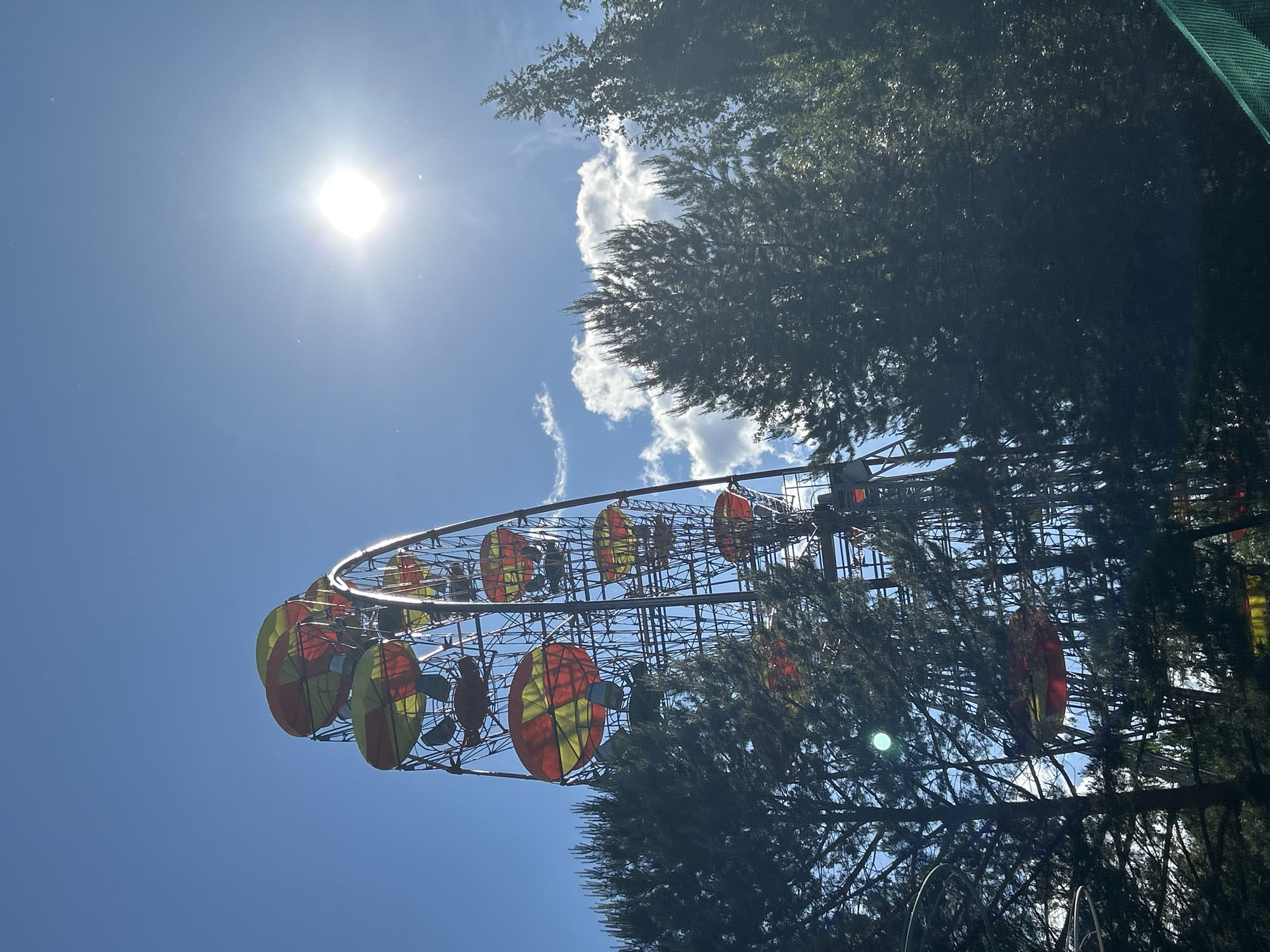 The image shows a decorative structure, possibly a Ferris wheel-like installation, adorned with red and yellow elements. The structure is situated outdoors, with a clear blue sky and bright sunlight visible in the background. Trees partially obscure the view of the structure. The location is likely Chaikovsky, Russia, but this cannot be determined from the image alone. No people are visible in the image.