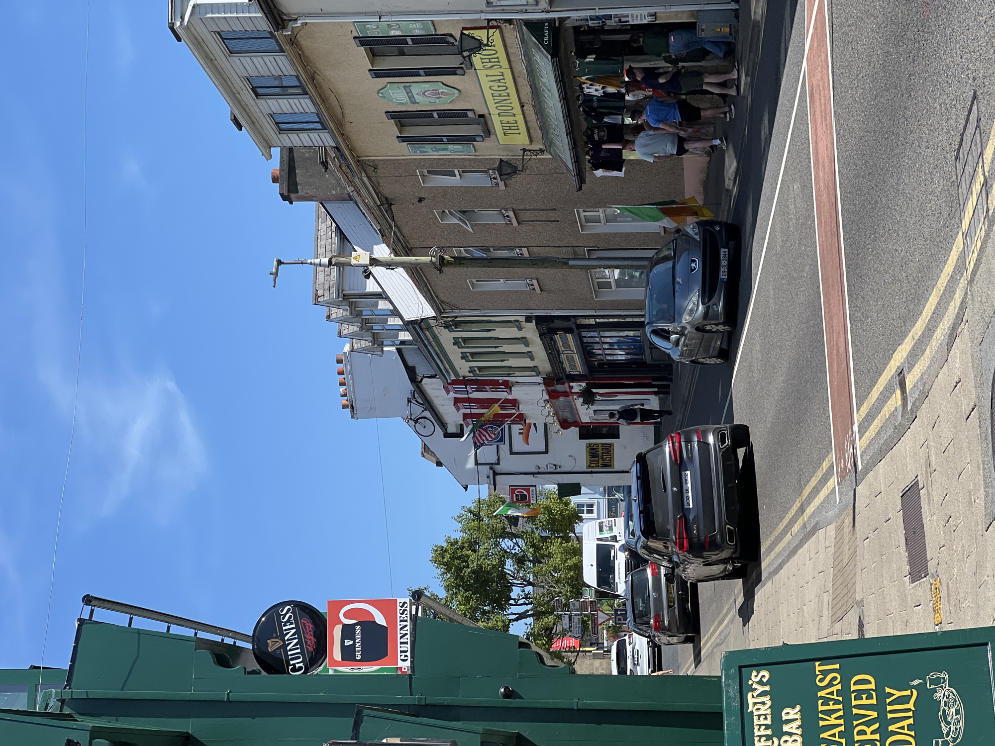 This is a street scene in Donegal Town, Ireland, on a sunny day. The camera is angled slightly upwards from a low vantage point. The foreground is dominated by a green sign with yellow lettering that reads "Rafferty's Bar," "Breakfast Served Daily." A crosswalk with red and white markings is visible on the asphalt street. Several cars are driving and parked on the street. On the left, a dark gray BMW SUV is in the foreground, followed by a lighter gray sedan. Further down the street, a white van is visible. On the right side of the street, there are several buildings with shops. One shop has a yellow sign that reads "The Donegal Shop" and displays clothing for sale outside. Other shops have red awnings and signage. People are seen walking on the sidewalk and entering shops. Two Irish flags are displayed on poles near the shops. The sky is a clear blue with some wispy clouds.