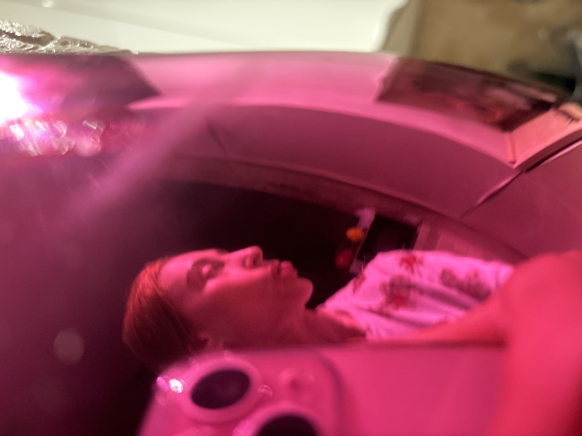 A blurred image, heavily tinted pink, shows a fair-skinned person lying supine inside what appears to be a CT scan machine. The person is wearing a light-colored, floral-patterned shirt. A portion of a cell phone is visible in the foreground. The location is not discernible from the image.